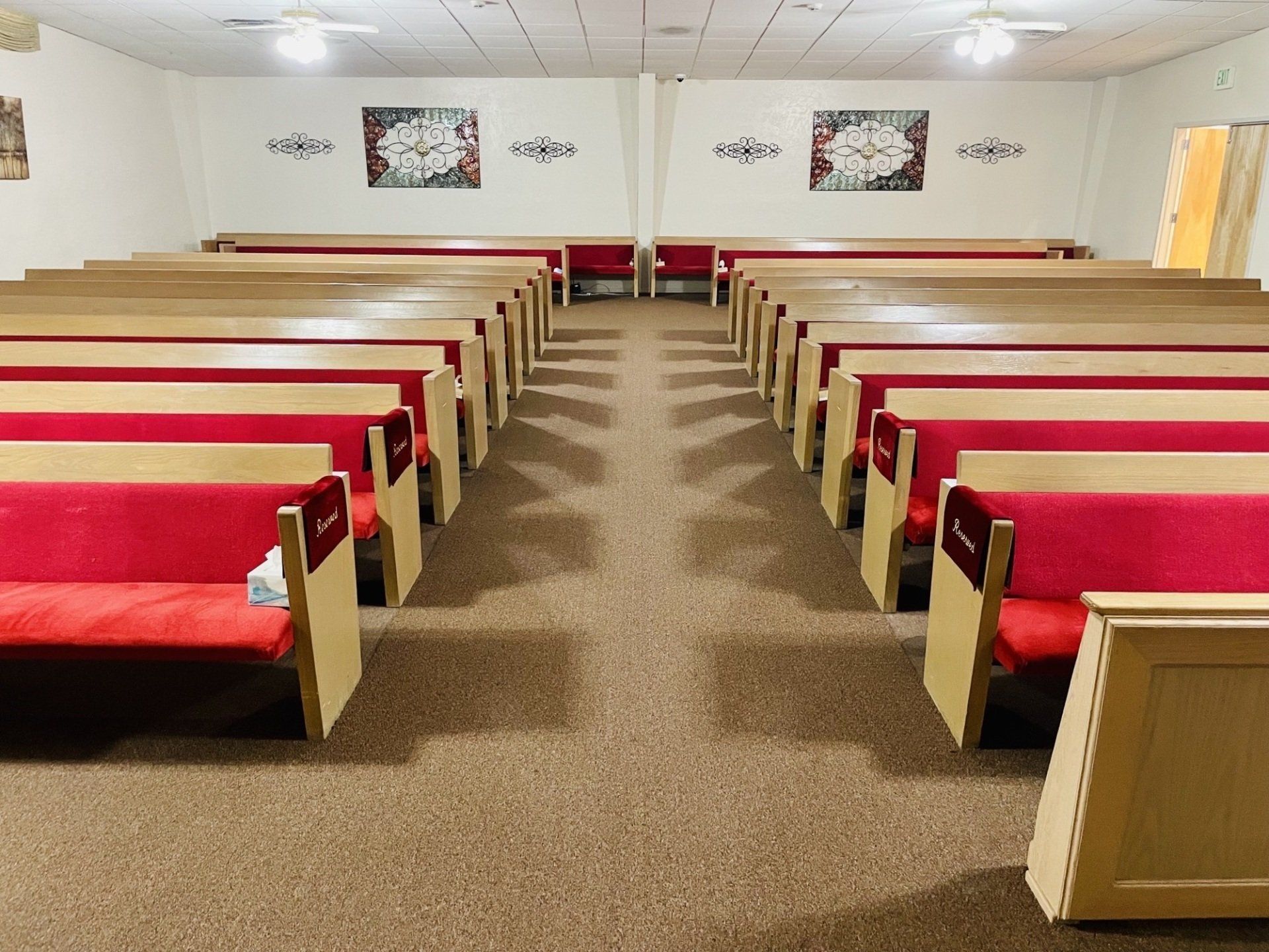 Rows of red-cushioned pews in a room with light beige walls and carpet, under ceiling lights.