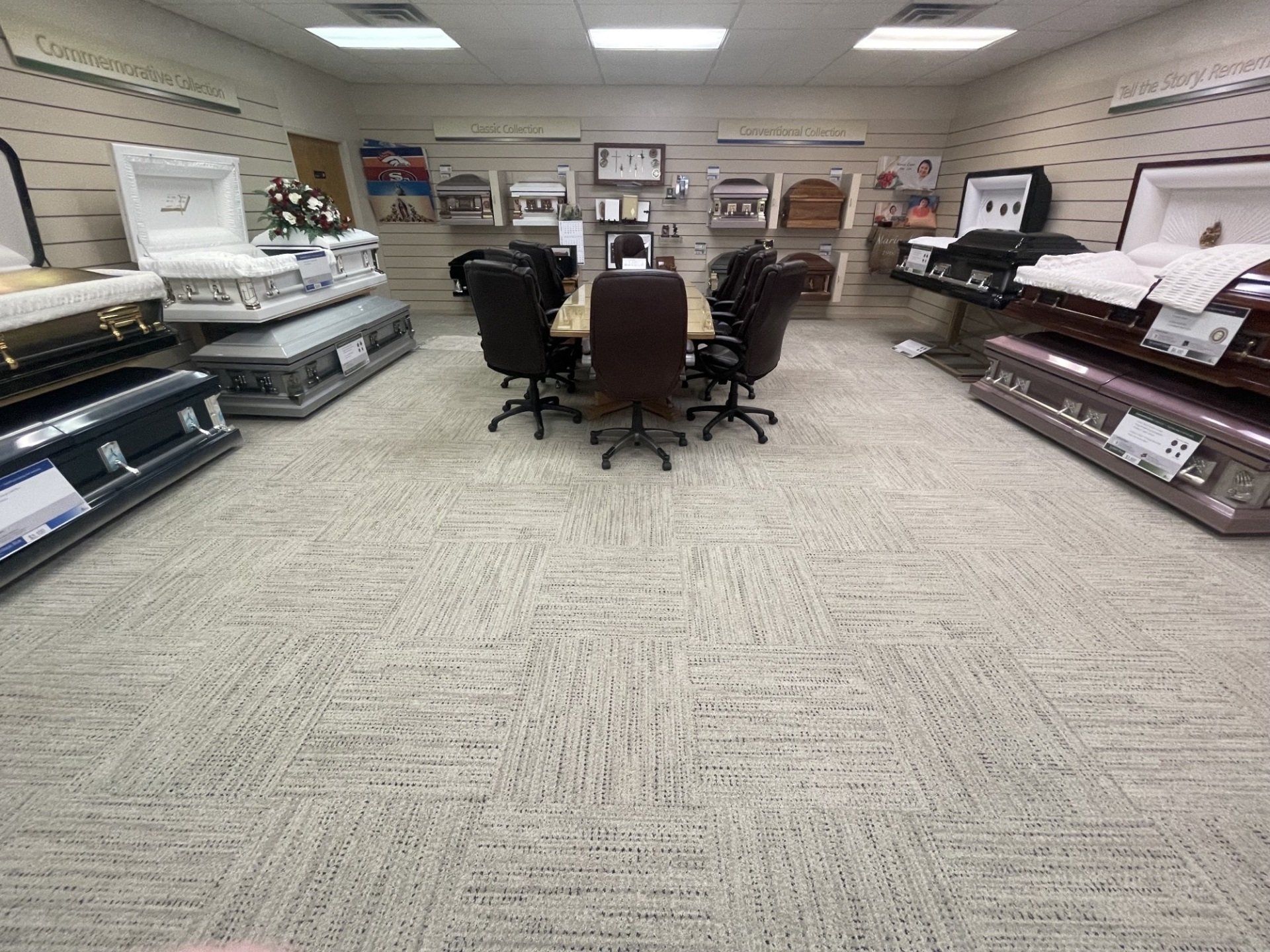 Interior of a funeral home showroom with caskets and a meeting table. Beige carpet and overhead lighting.