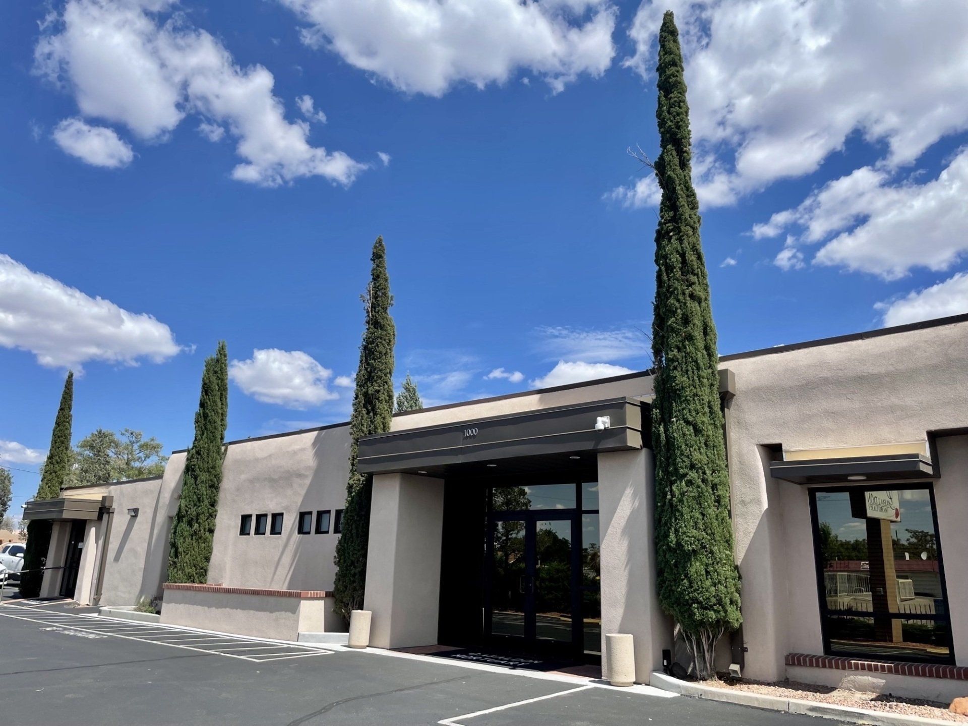 A beige building with tall, slender trees, under a blue sky with fluffy clouds.