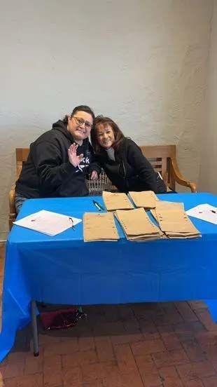 Two people smile behind a blue table with papers, one waving.