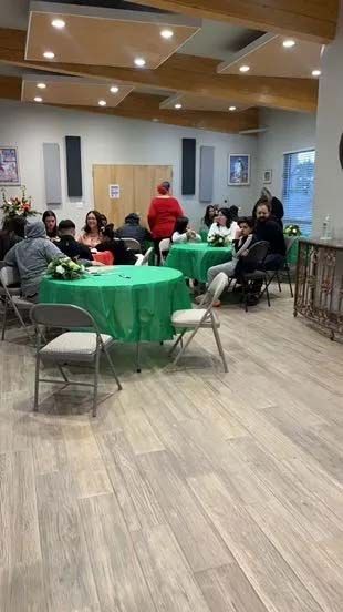 People seated at green-covered tables in a room with speakers and wood-look flooring. A woman in red stands.