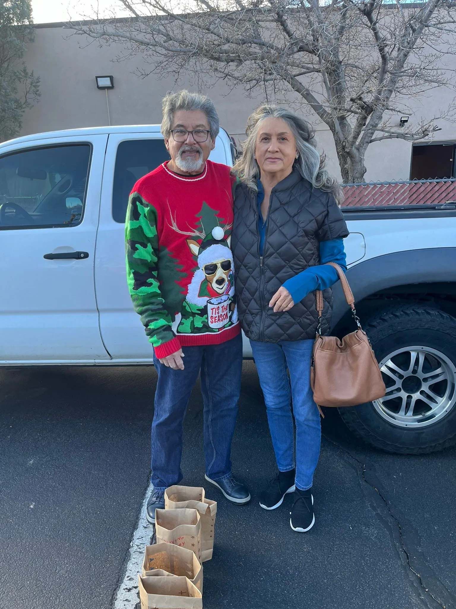 Couple standing near a truck; man in a Christmas sweater, woman in a vest, near paper bags.