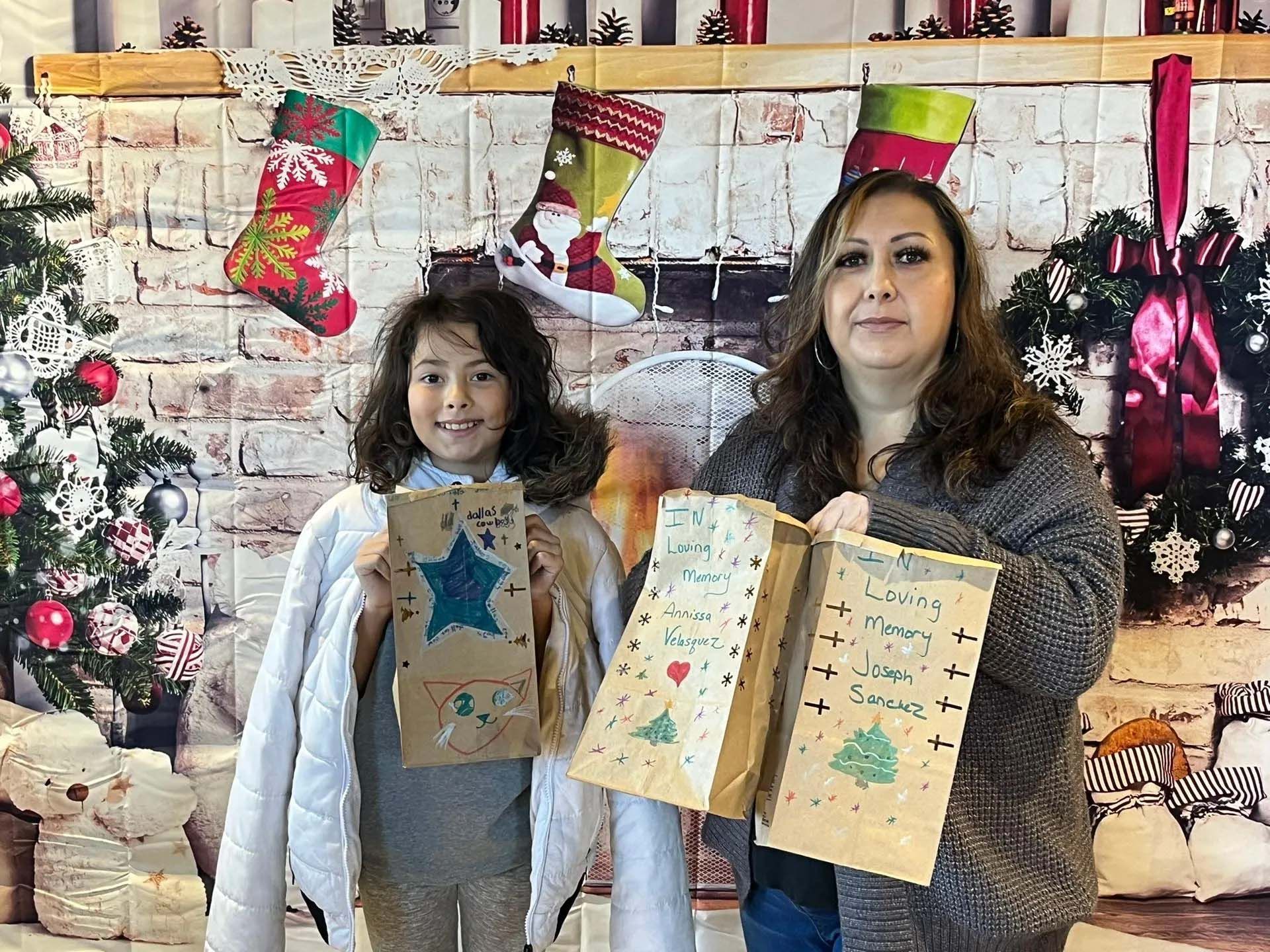 Woman and child holding decorated paper bags in front of a Christmas-themed backdrop with stockings and a tree.