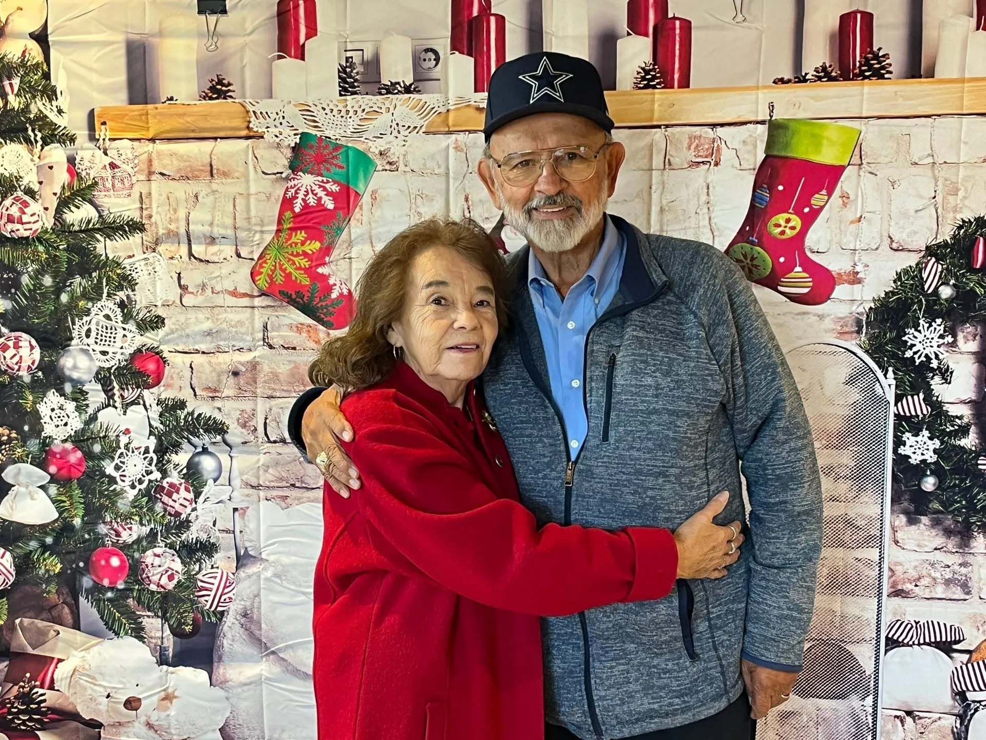 Couple hugging in front of a Christmas backdrop with a tree and stockings.