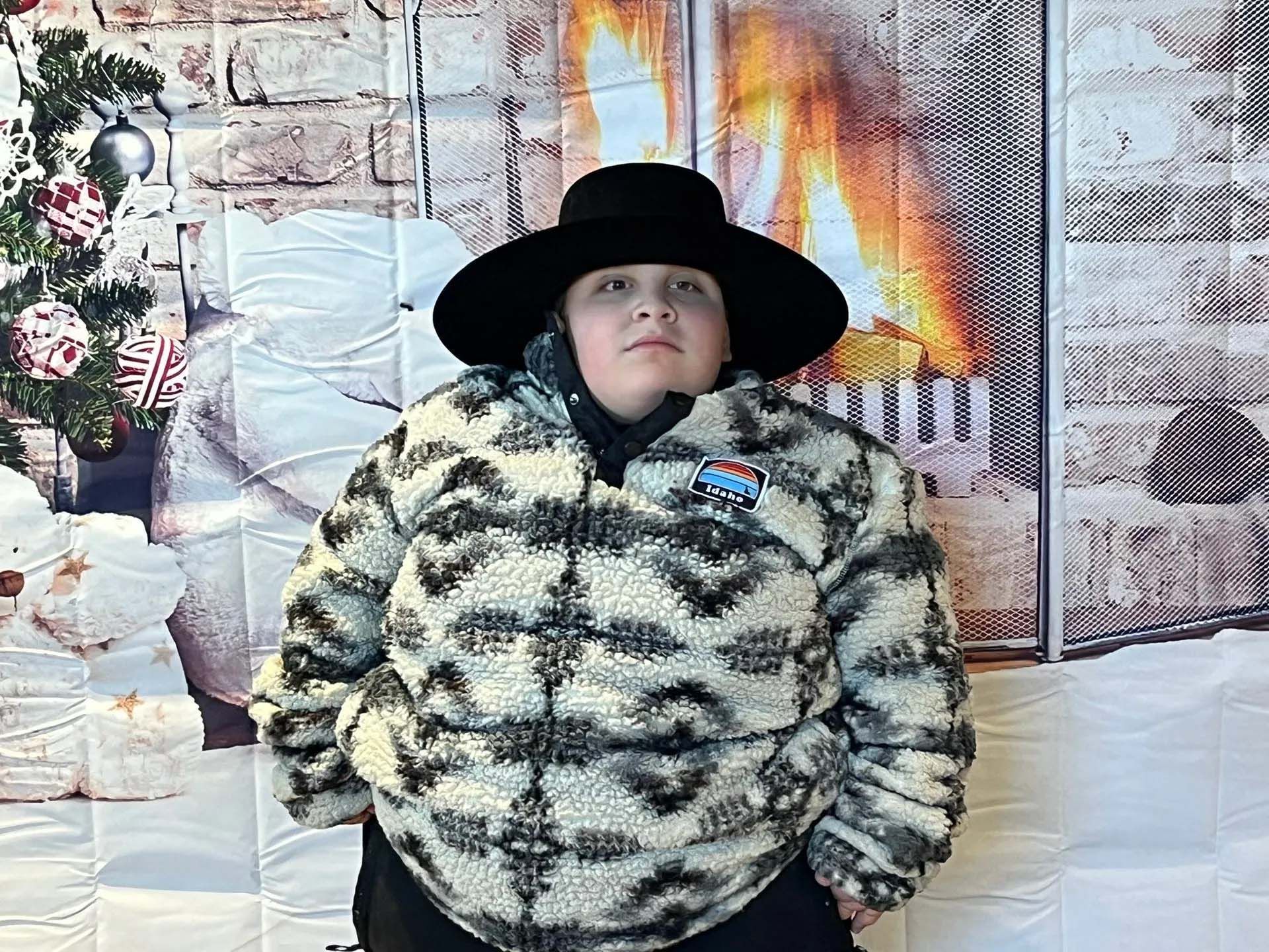 Person wearing a wide-brimmed black hat and a patterned fleece jacket, standing in front of a fireplace and Christmas decorations.