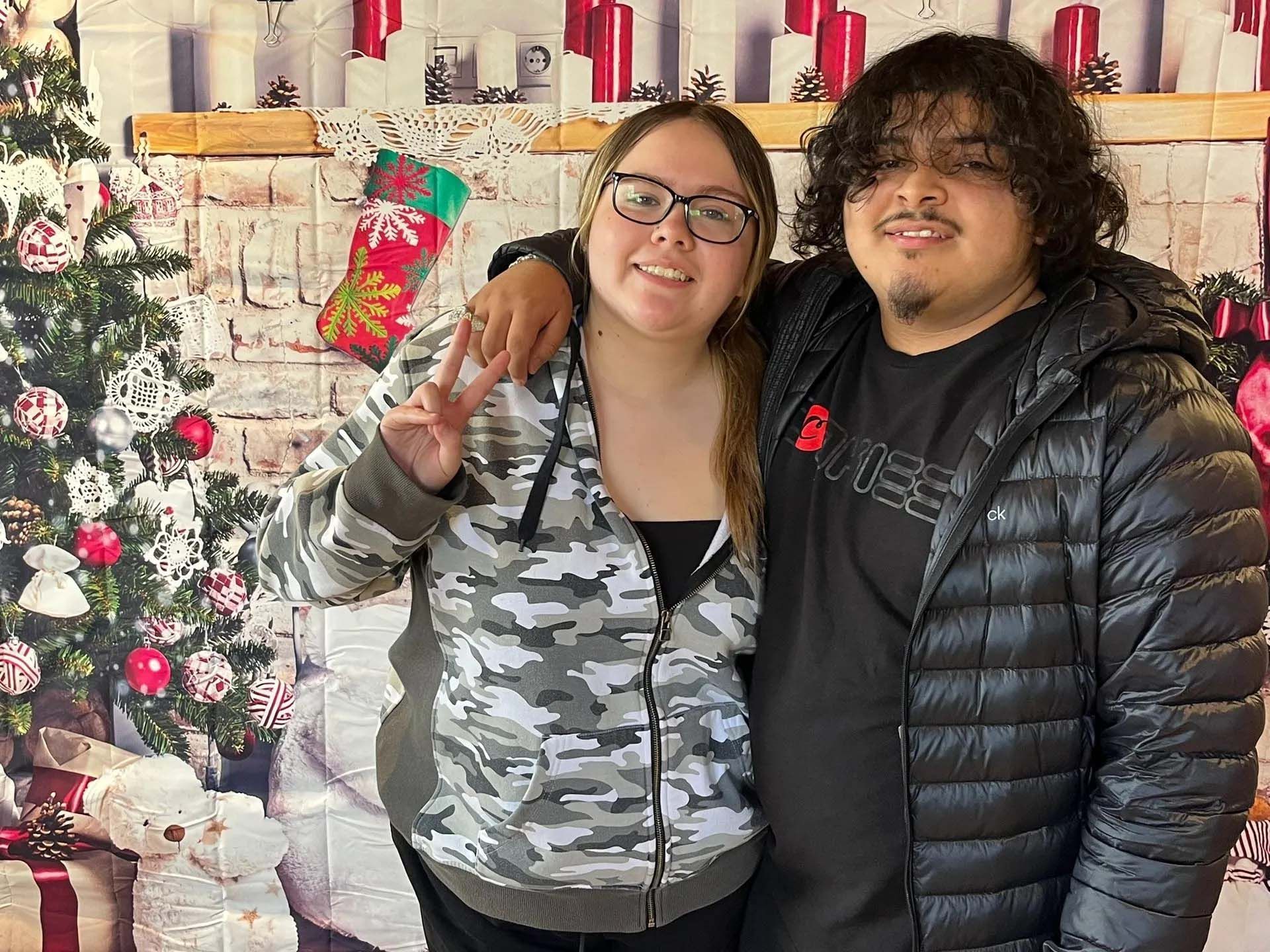 Two people posing by a Christmas backdrop; one person gives a peace sign, both smiling.