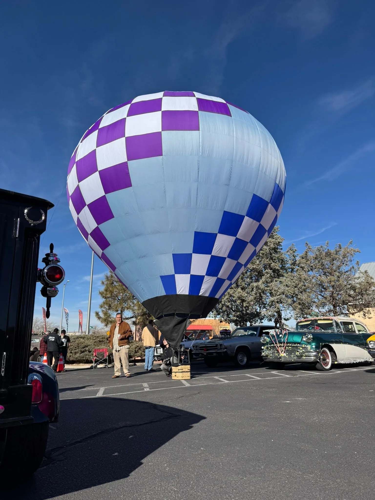 Hot air balloon with purple and white checkered pattern, over cars in parking lot.