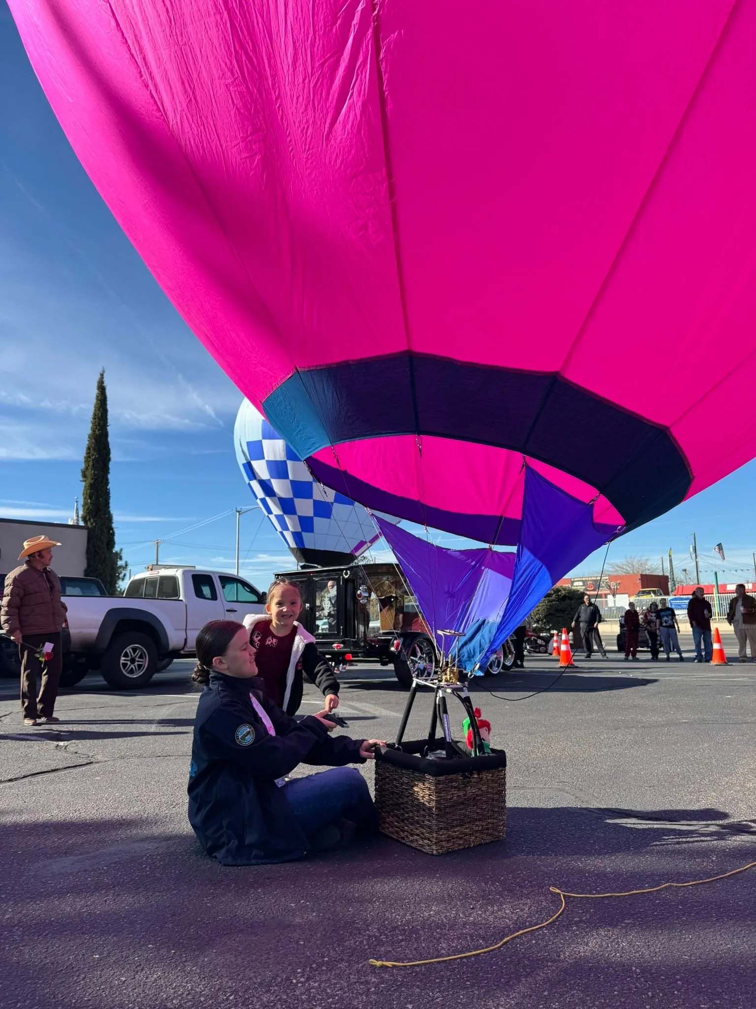 Person preparing hot air balloon for takeoff in a parking lot. Pink and blue balloon, blue sky.