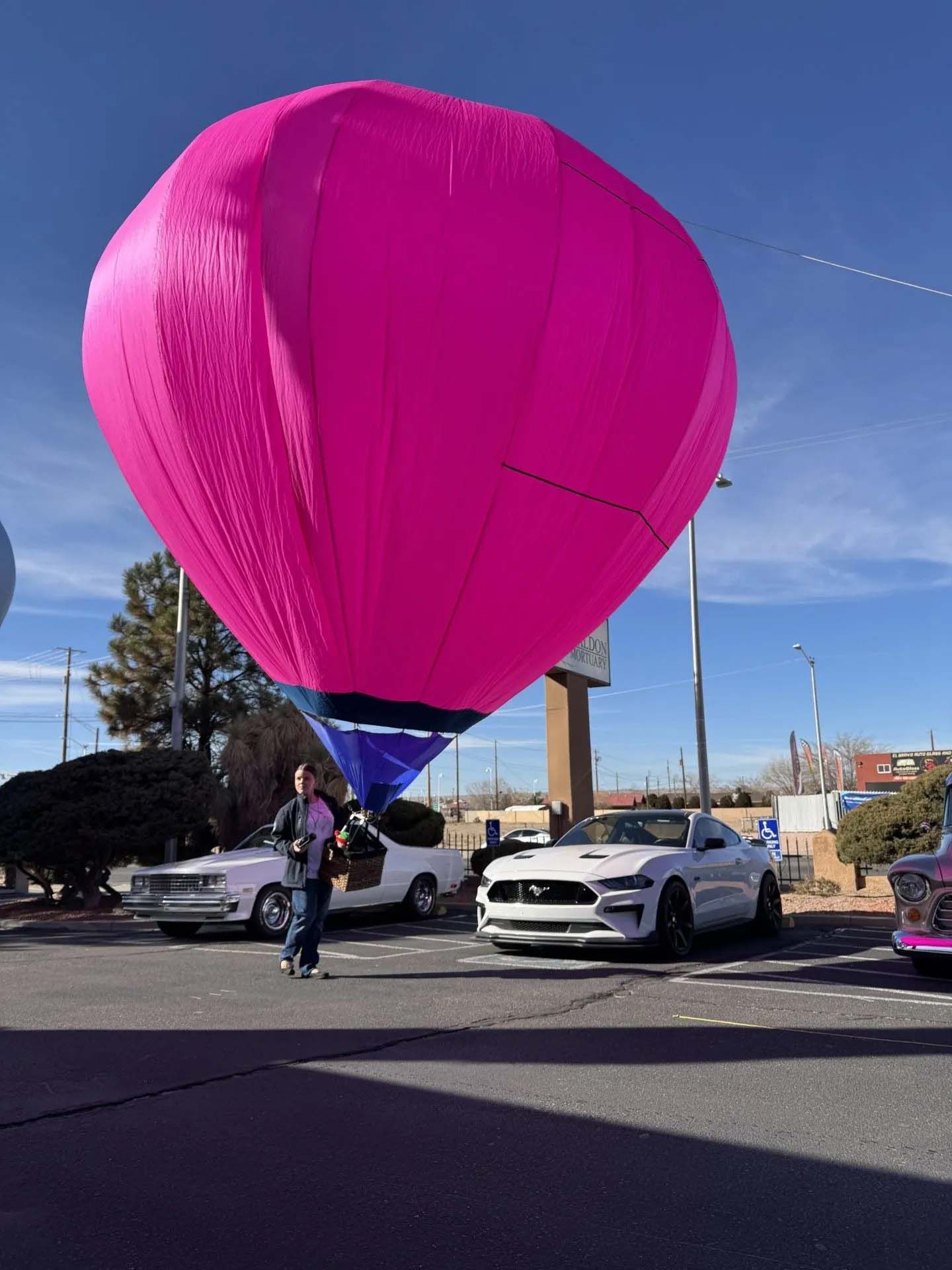 Pink hot air balloon over parked cars, a person stands nearby. Bright blue sky.