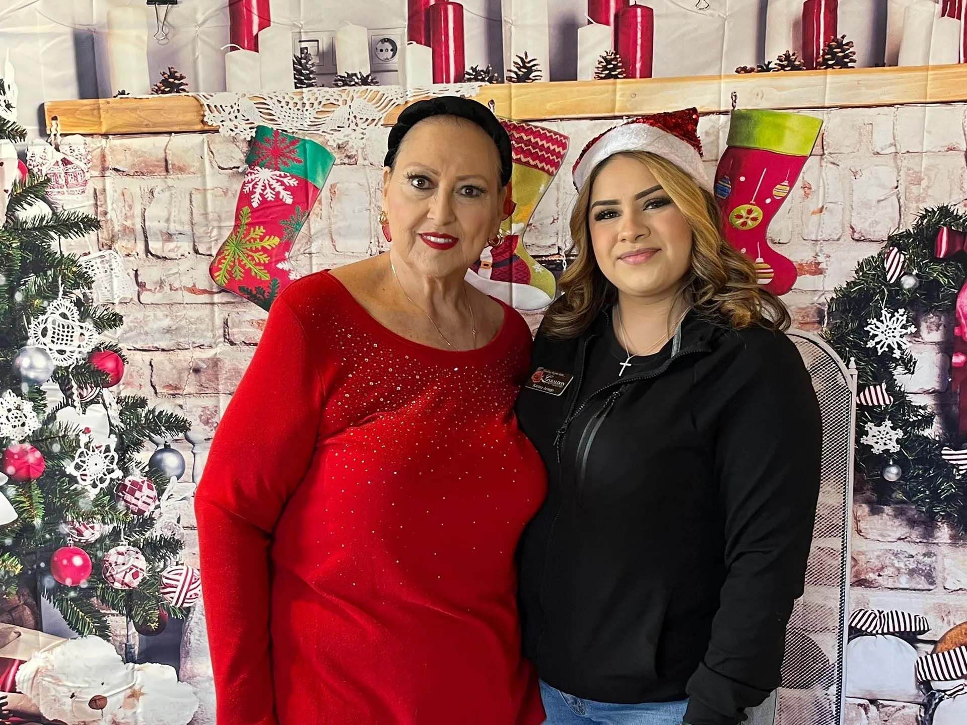 Woman in red sweater and another with Santa hat posing in front of a Christmas backdrop.