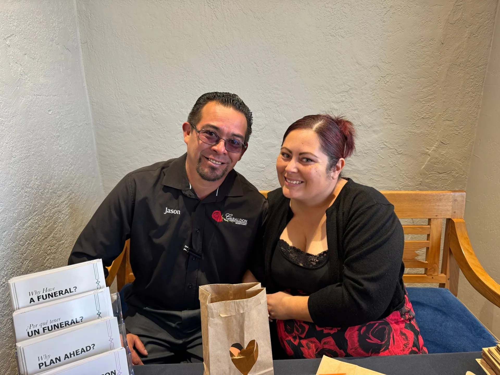 Man and woman smiling behind a table with boxes and a paper bag with a heart cutout; indoor setting.