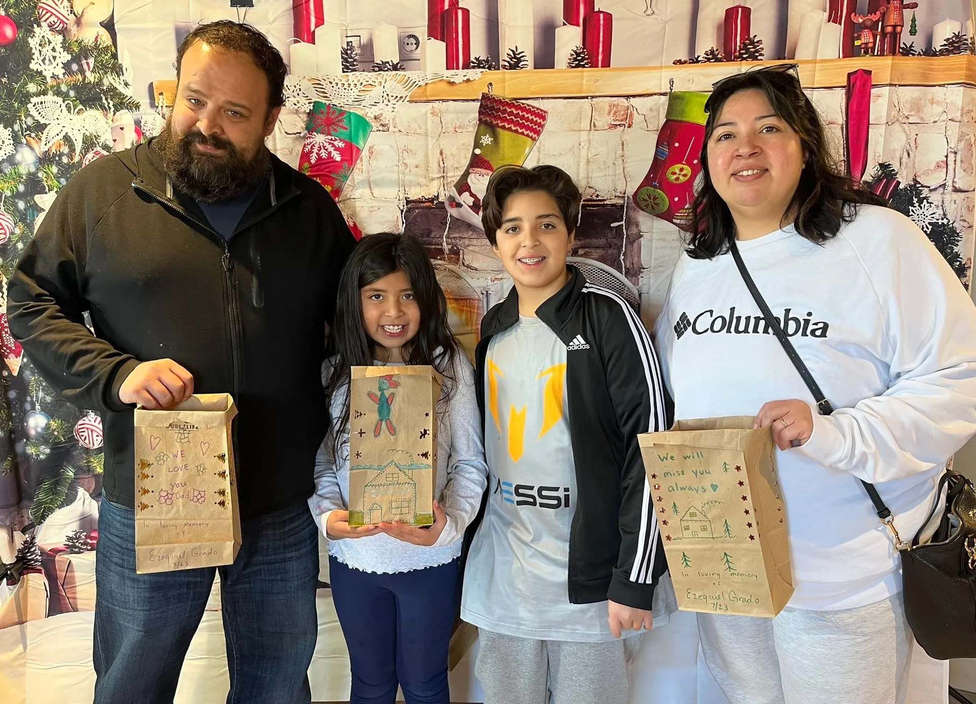 Family smiling, holding decorated paper bags, in front of Christmas decorations.
