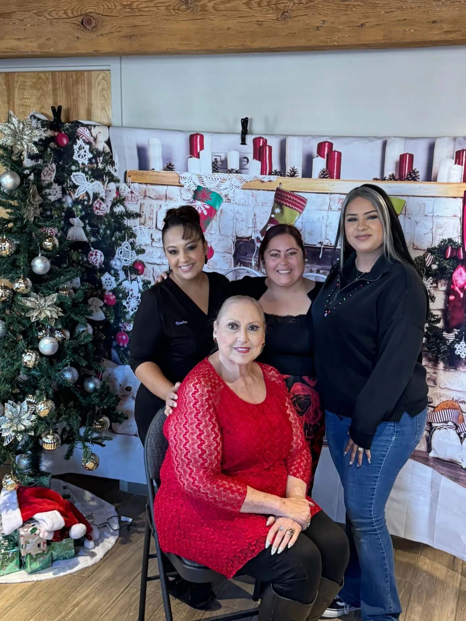Four people pose for a photo near a decorated Christmas tree and festive background. Woman in red dress sits.