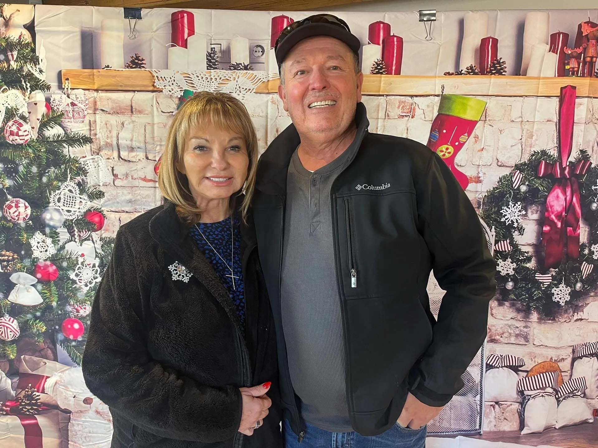 Couple posing in front of a Christmas-themed backdrop. Woman in black coat, man in jacket and cap, both smiling.