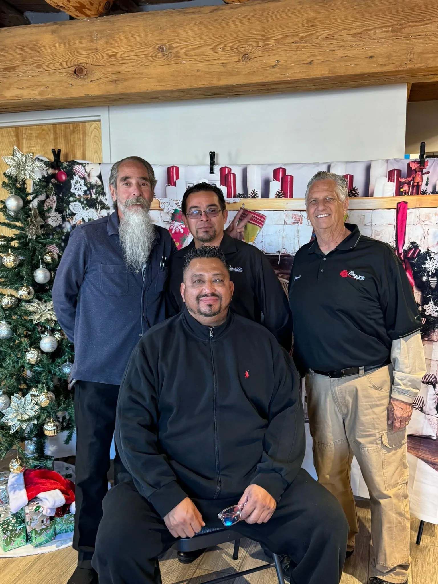 Four men standing in front of a Christmas tree and decorated wall. One man is seated in front.