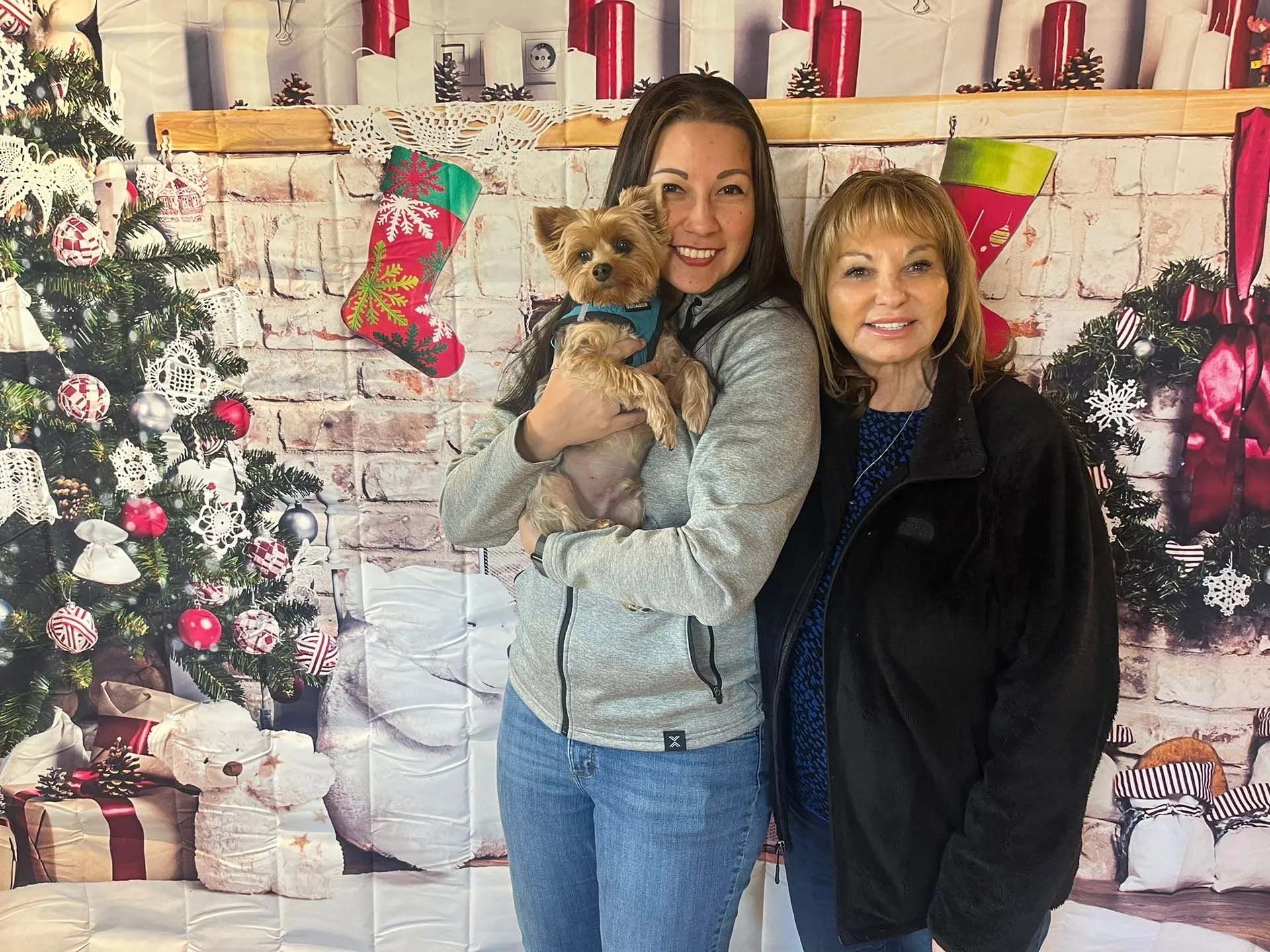 Two women and small dog pose in front of a Christmas backdrop.