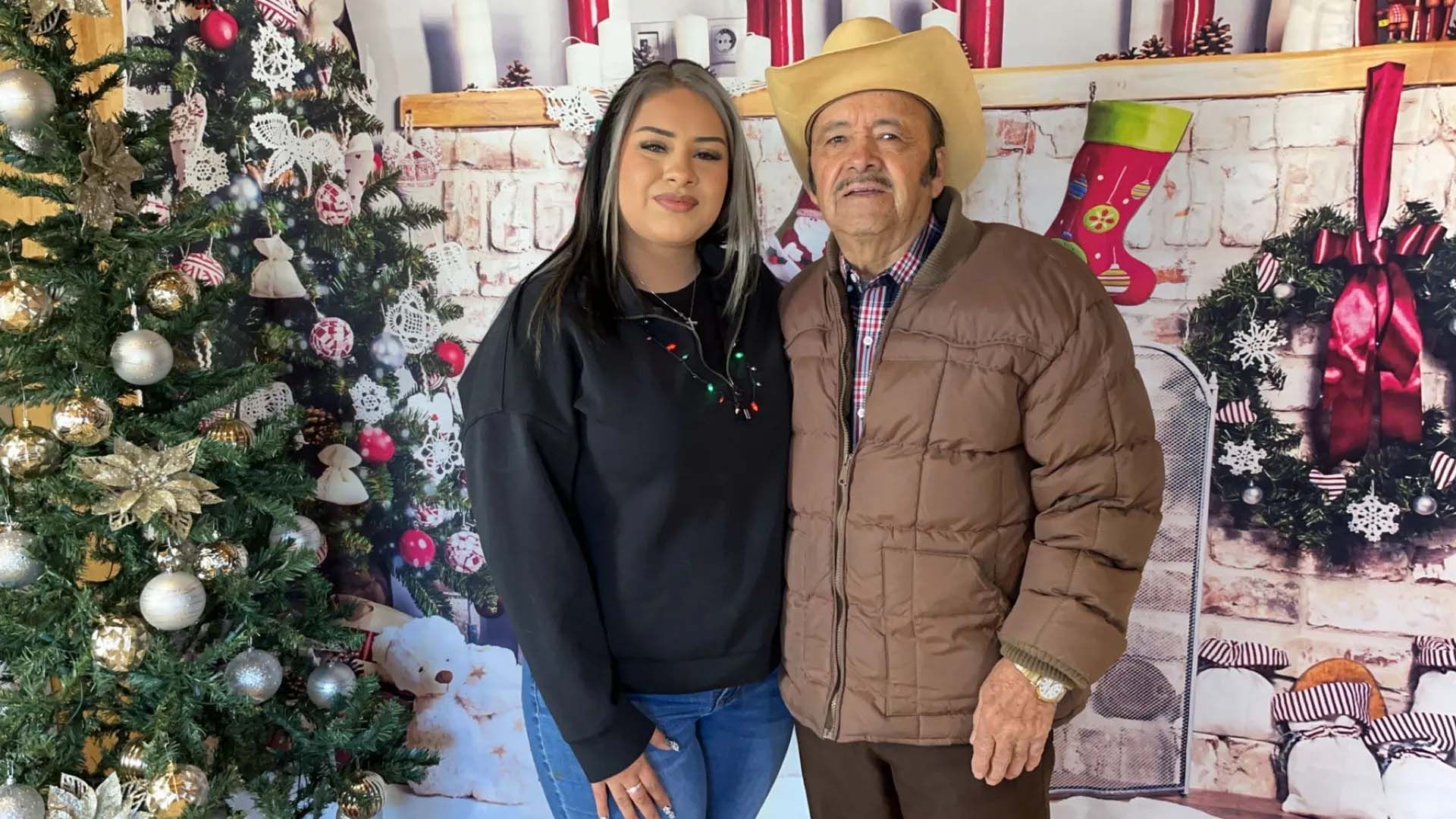 Woman and man stand in front of a Christmas-themed backdrop. Man wears a hat and brown jacket.