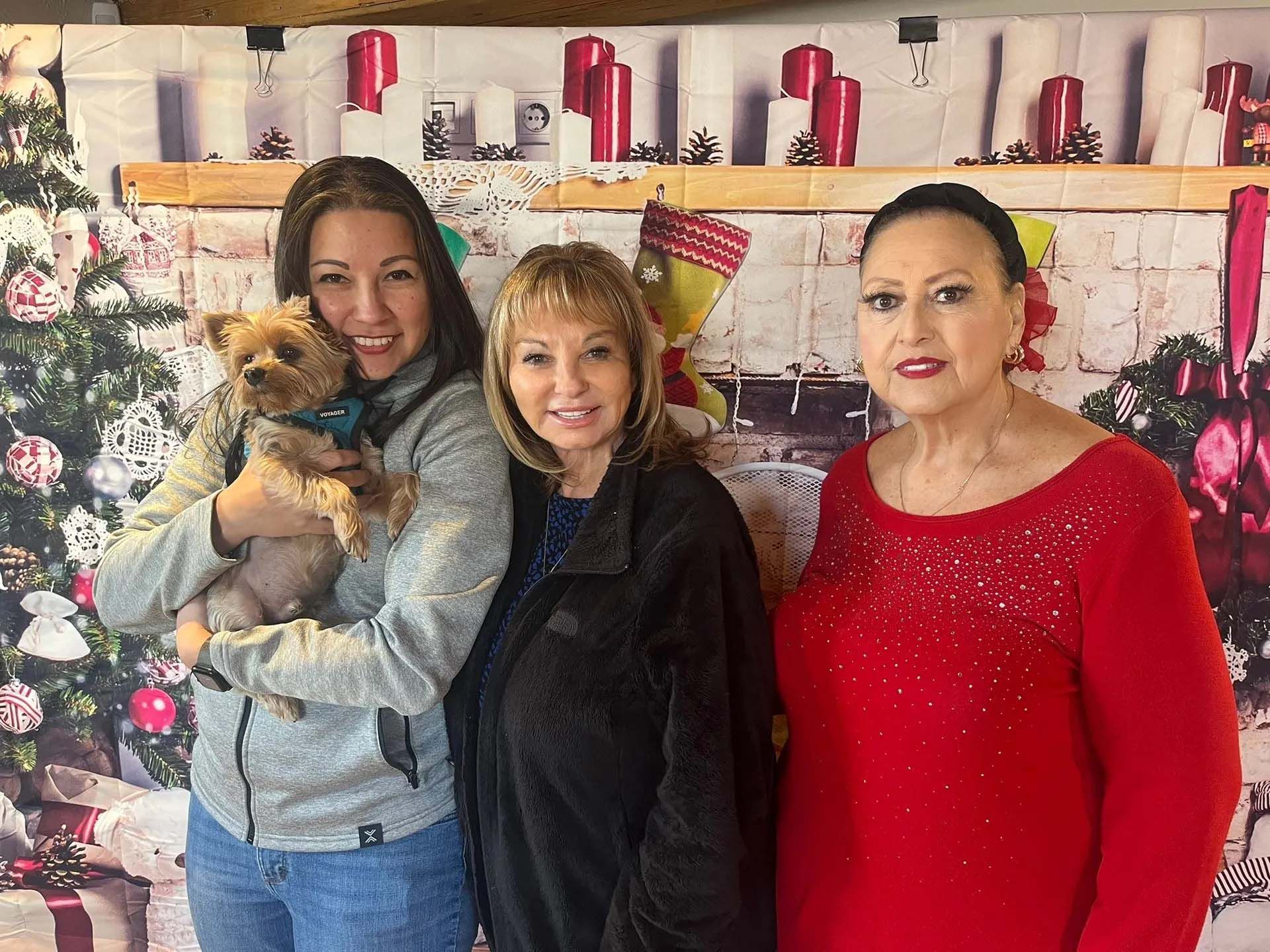 Three women and a dog pose in front of a festive backdrop. One woman holds the dog, another wears a red sweater.