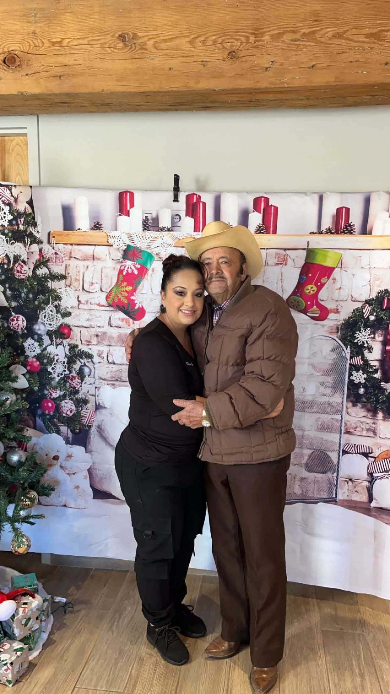 Woman and man in front of a Christmas backdrop. They are embracing. The man wears a cowboy hat.