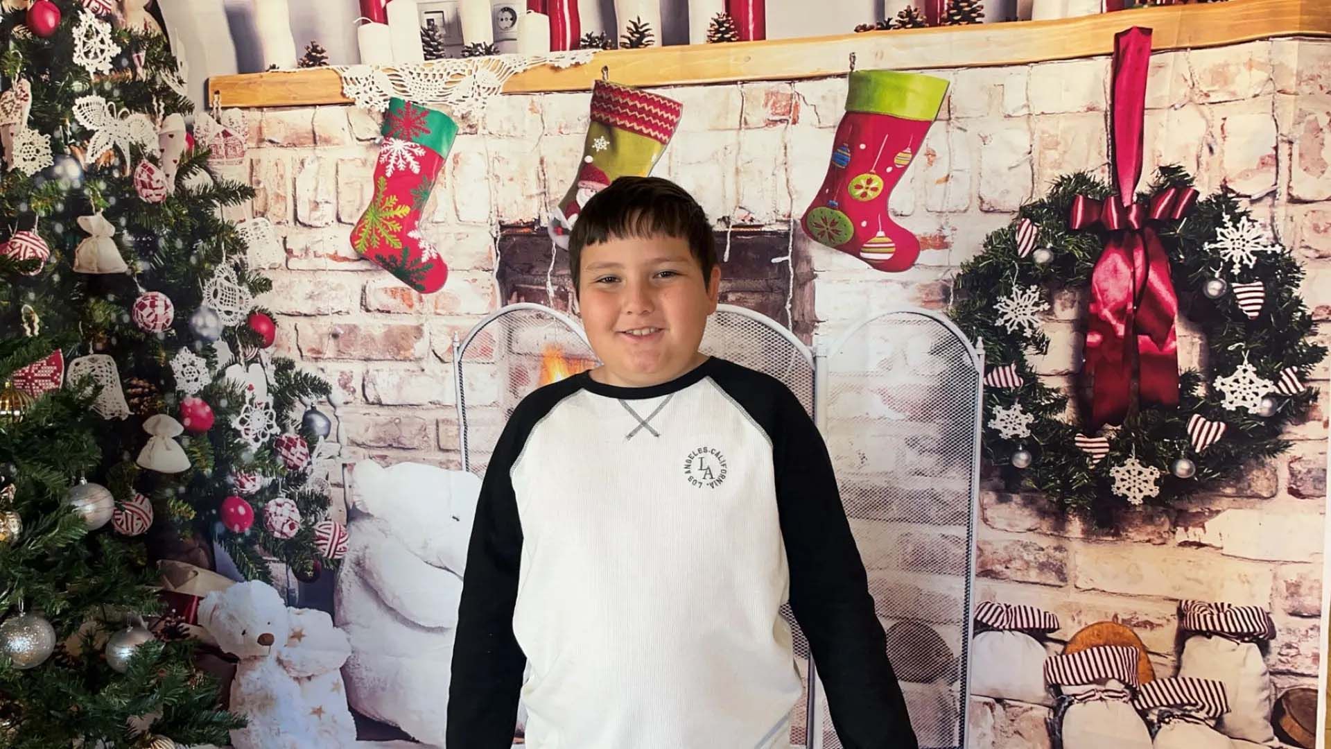 Boy in black and white shirt smiles in front of Christmas backdrop with stockings, wreath, and tree.