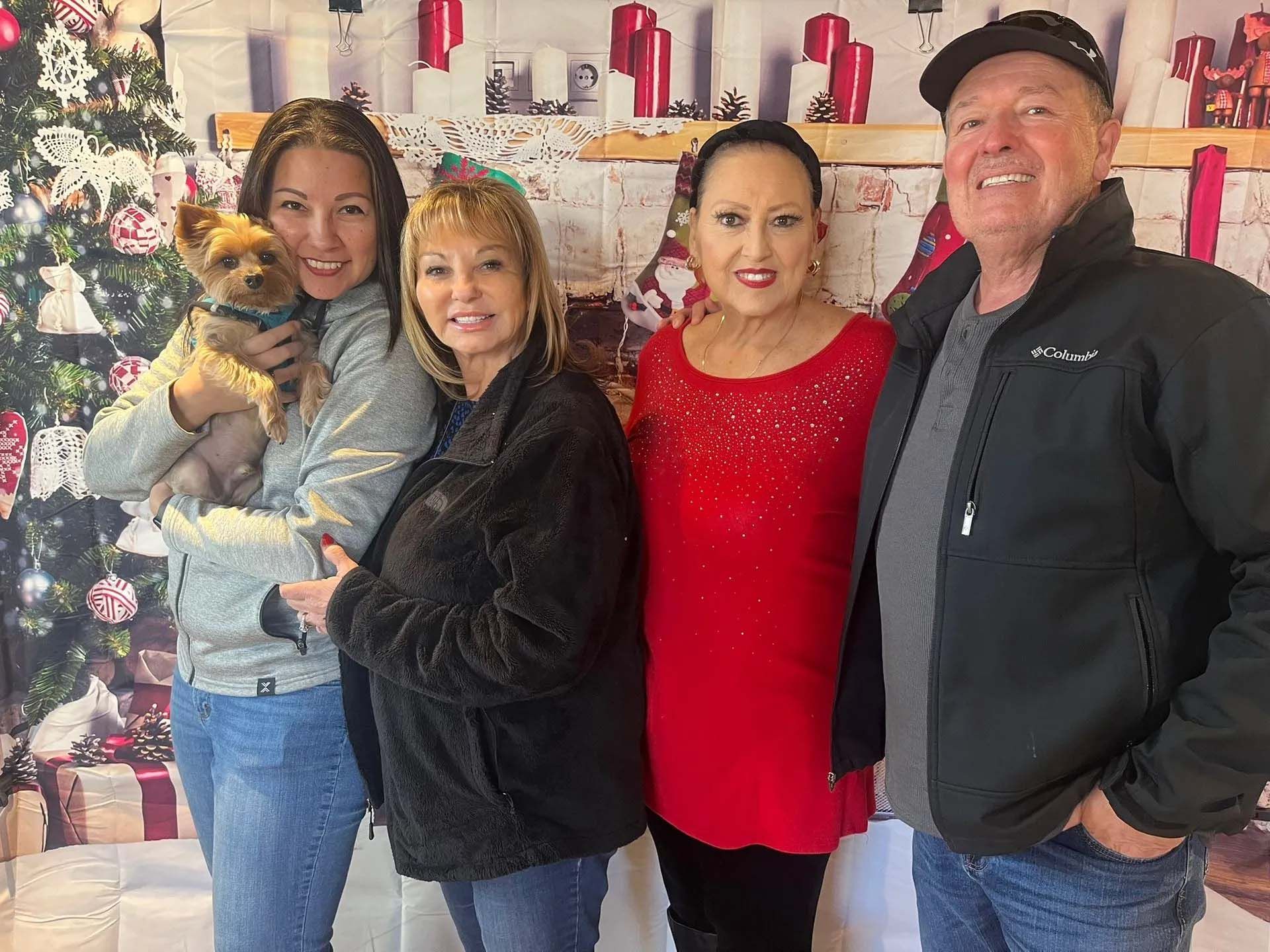 Four people smiling, posing in front of a Christmas backdrop. Woman holds small dog.
