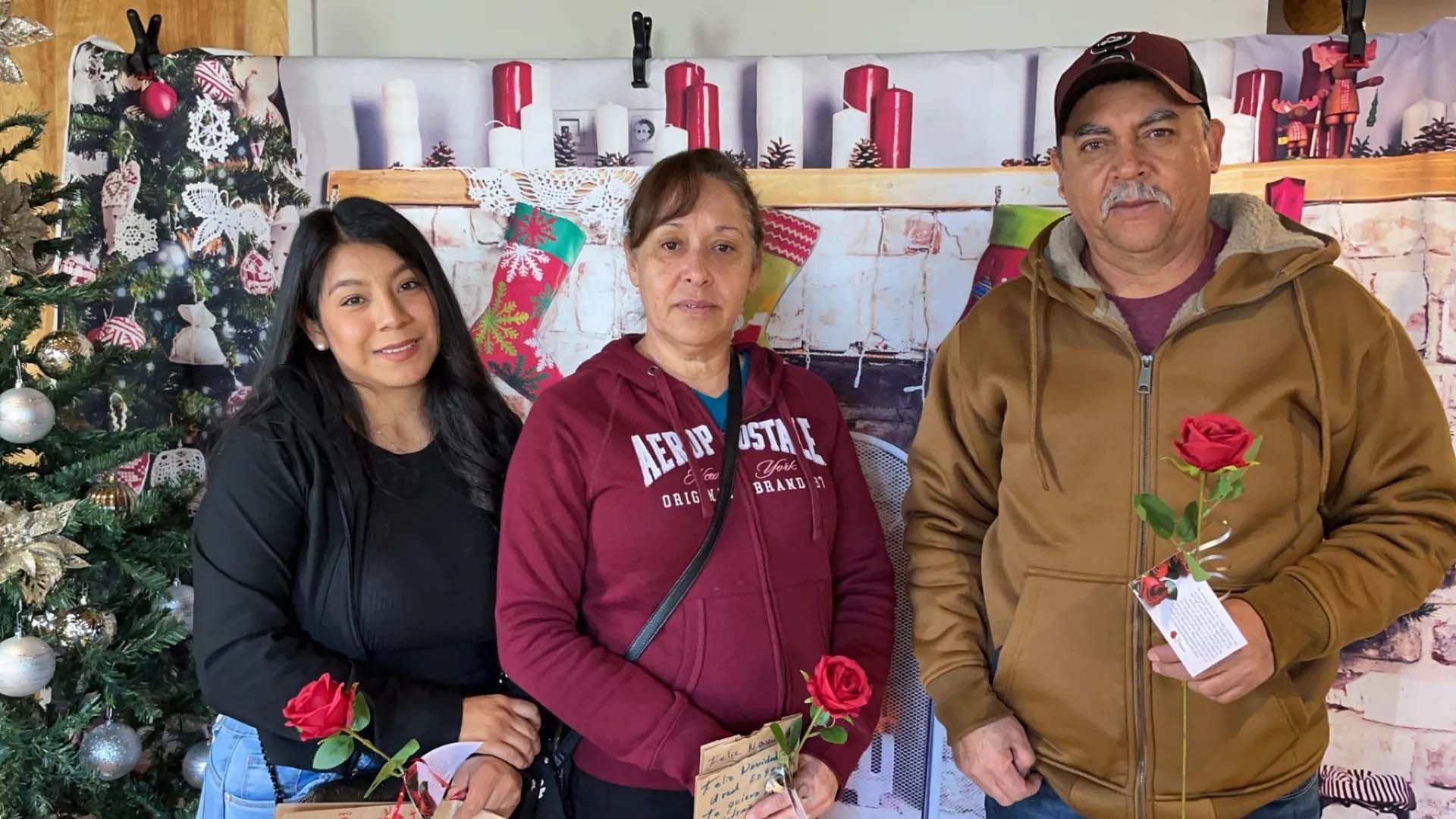 Three people holding red roses pose in front of a Christmas backdrop.