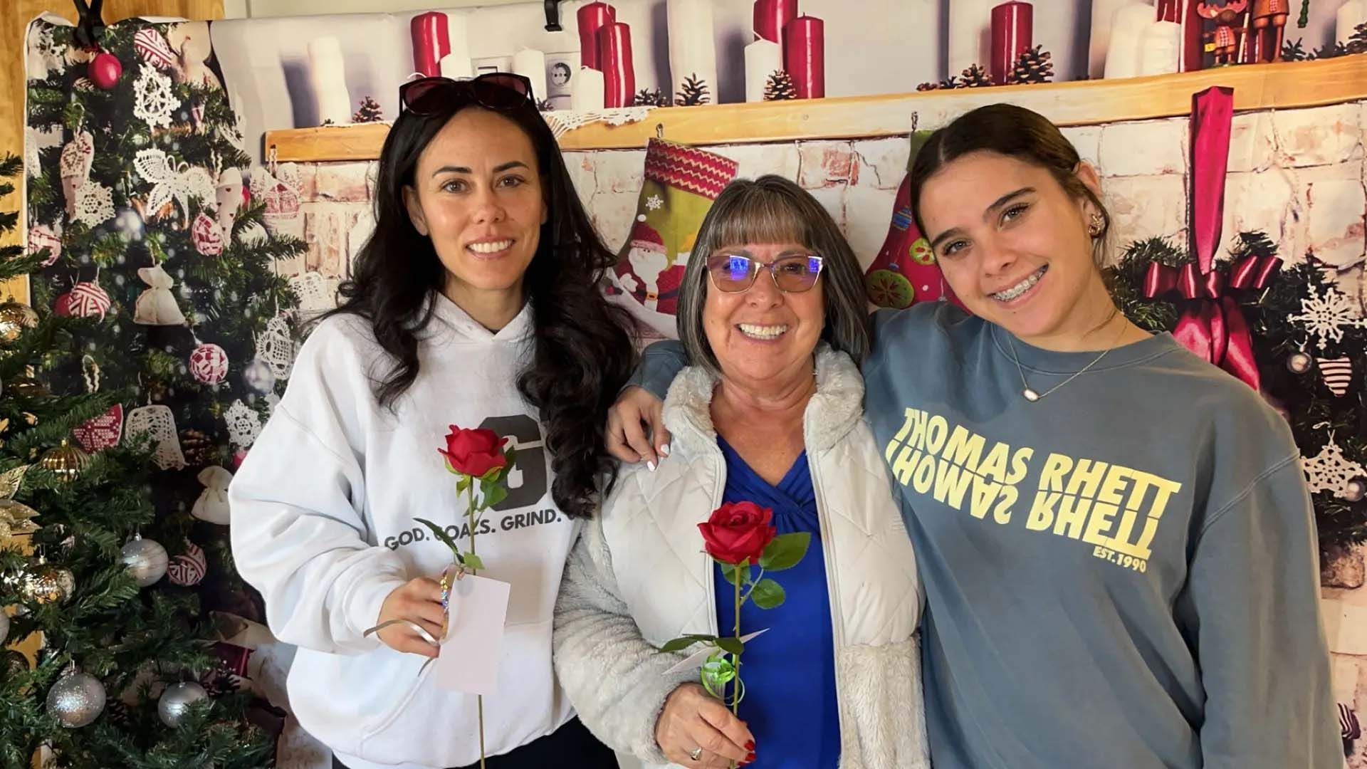 Three women holding red roses, smiling, posing in front of Christmas decorations.