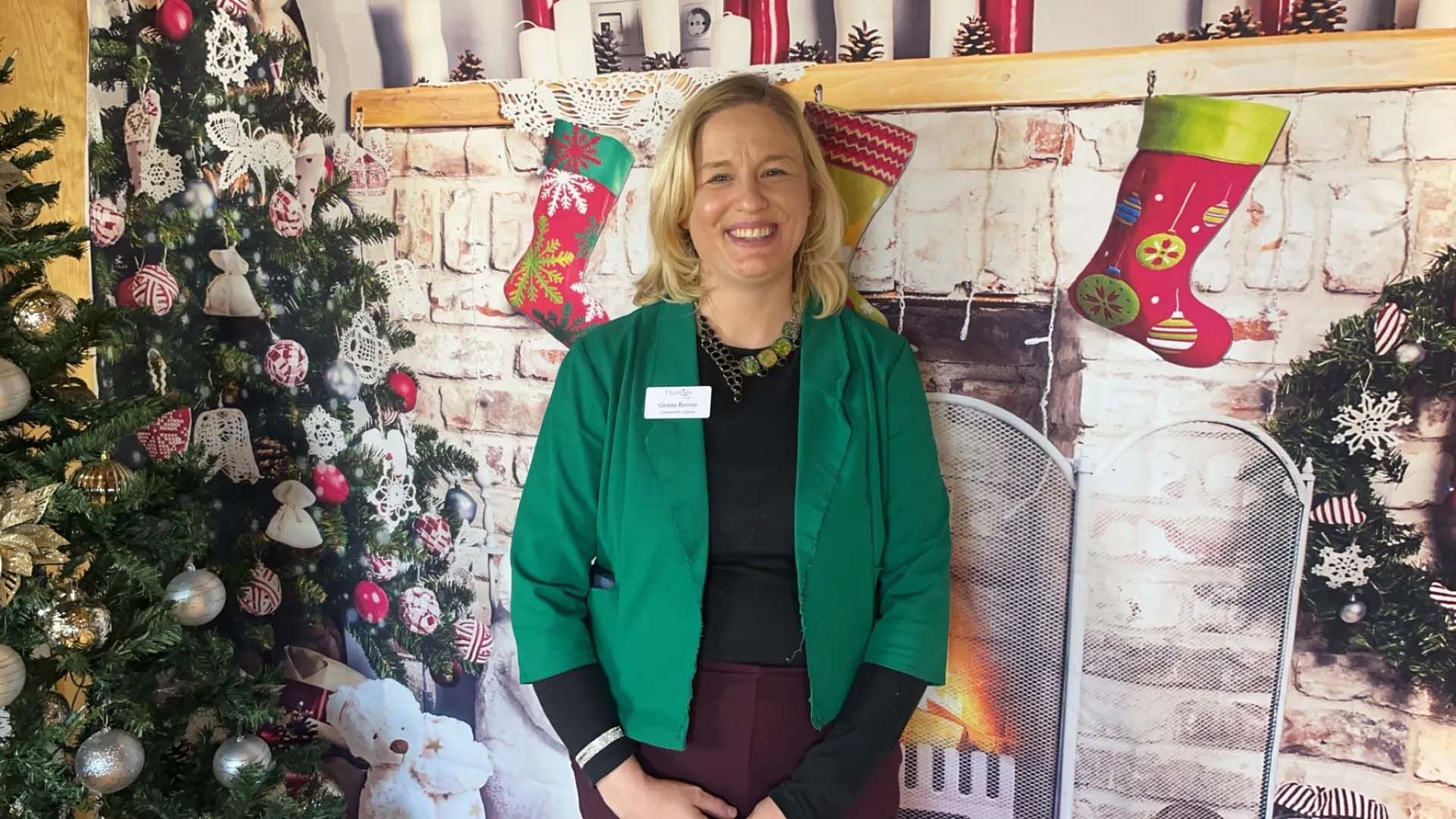 Woman in green jacket stands in front of Christmas decorations, smiling.