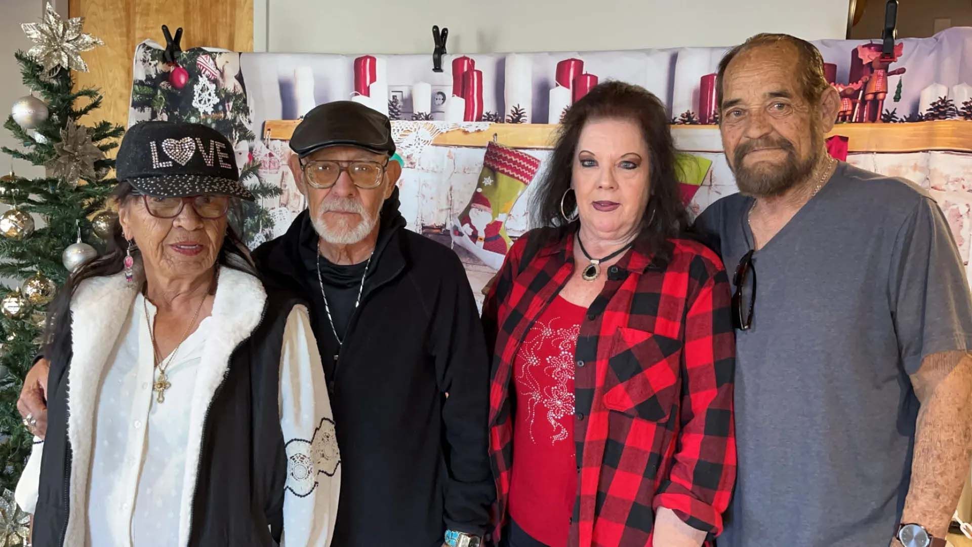 Four people standing indoors; woman wearing hat, woman in plaid, two men, Christmas decorations in the background.