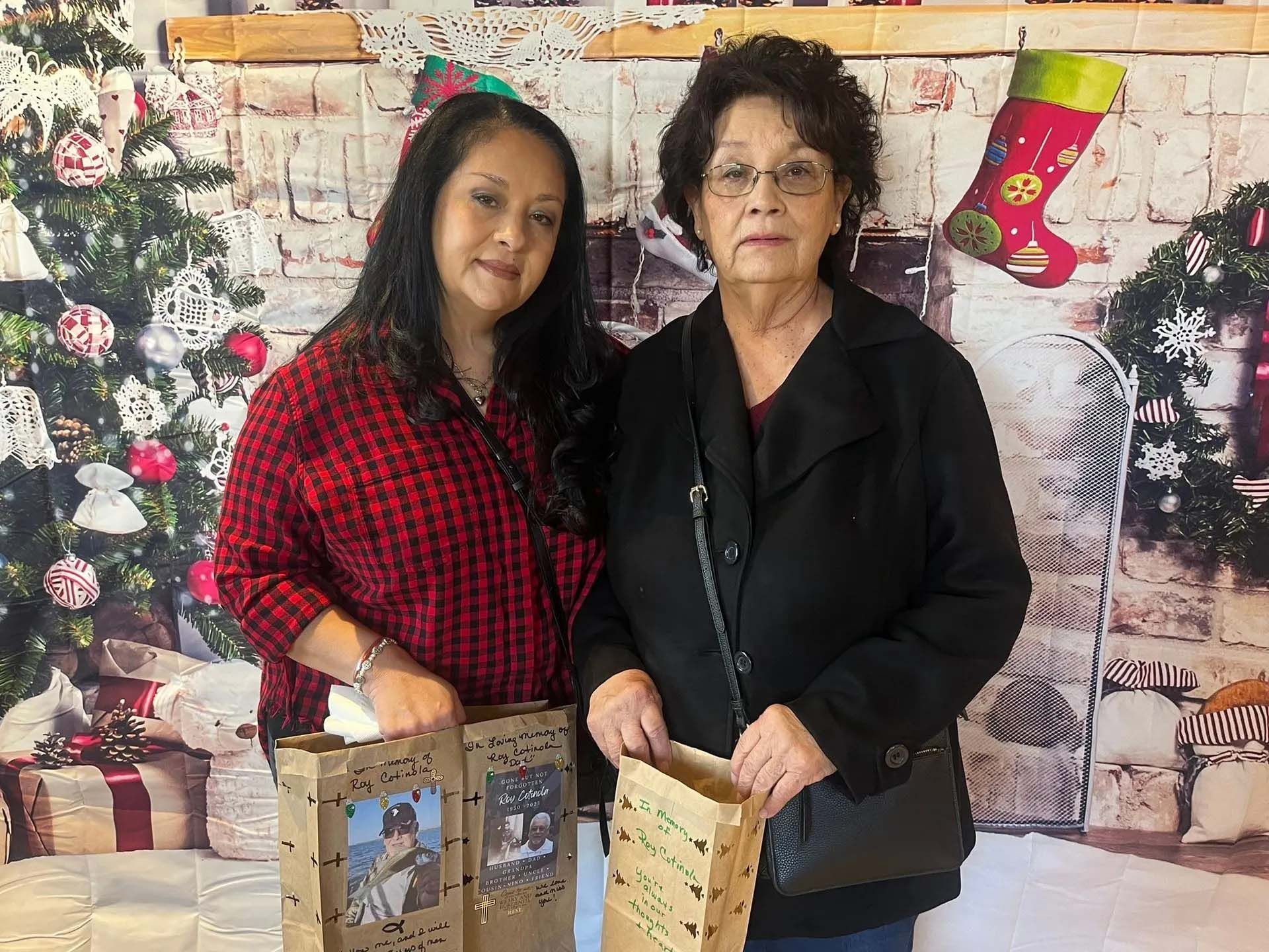 Two women stand by a table with gift bags, in front of a Christmas-themed backdrop.