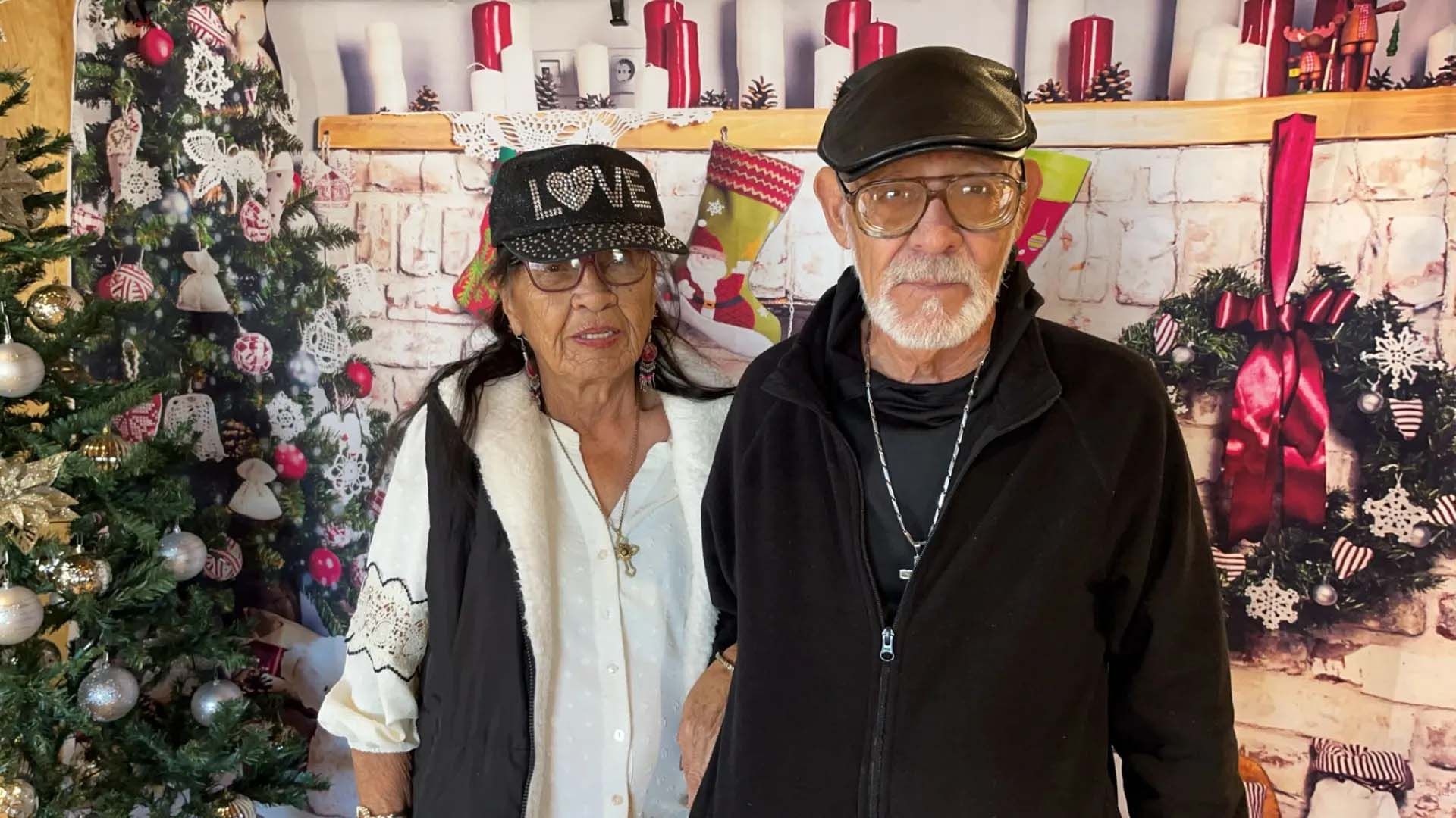 Couple standing in front of a Christmas-themed backdrop. They smile at the camera.