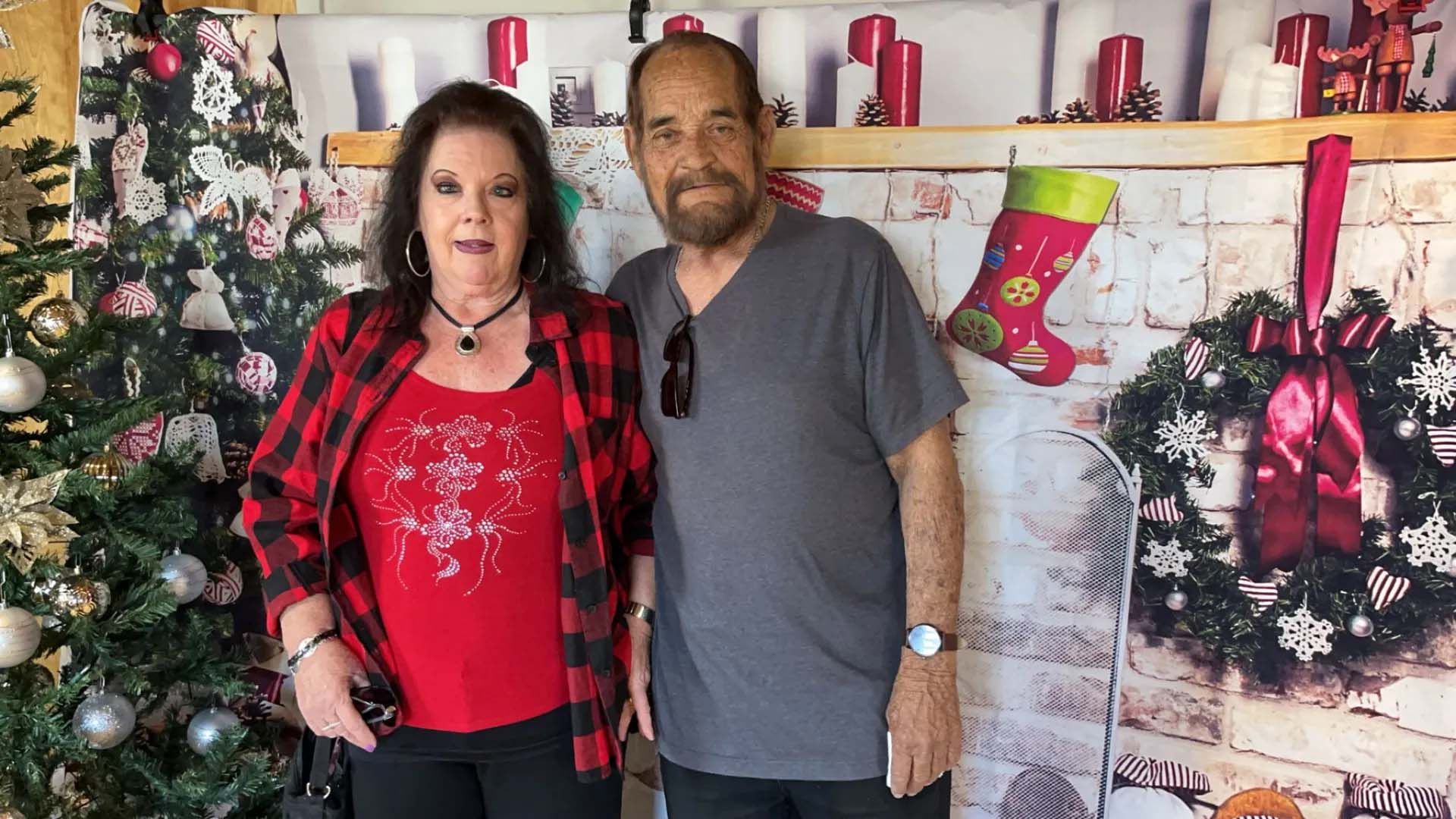 Couple posing in front of a Christmas backdrop. Woman wears a red plaid shirt, man in gray.