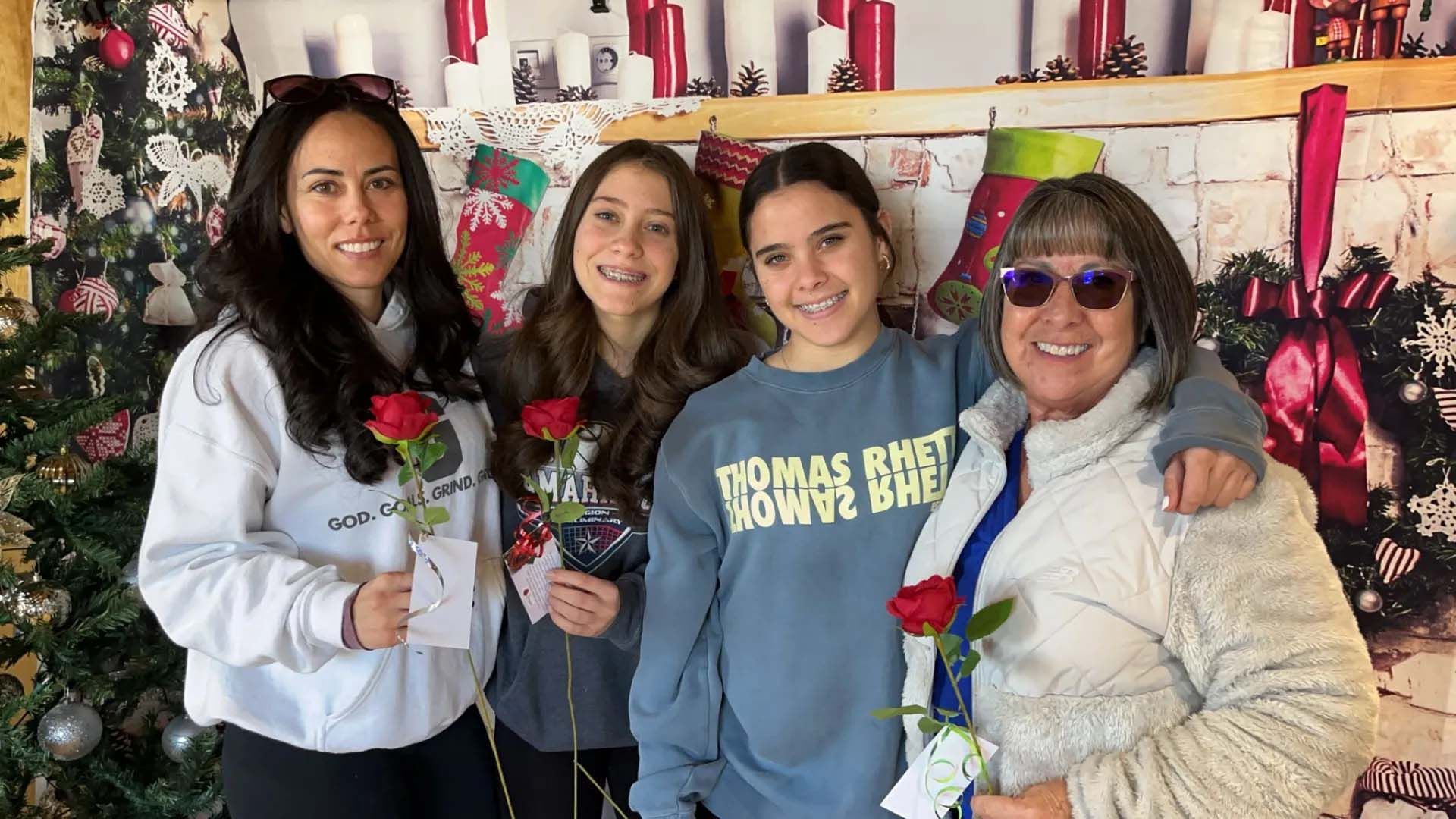 Four people posing, holding roses in front of a Christmas-themed backdrop.