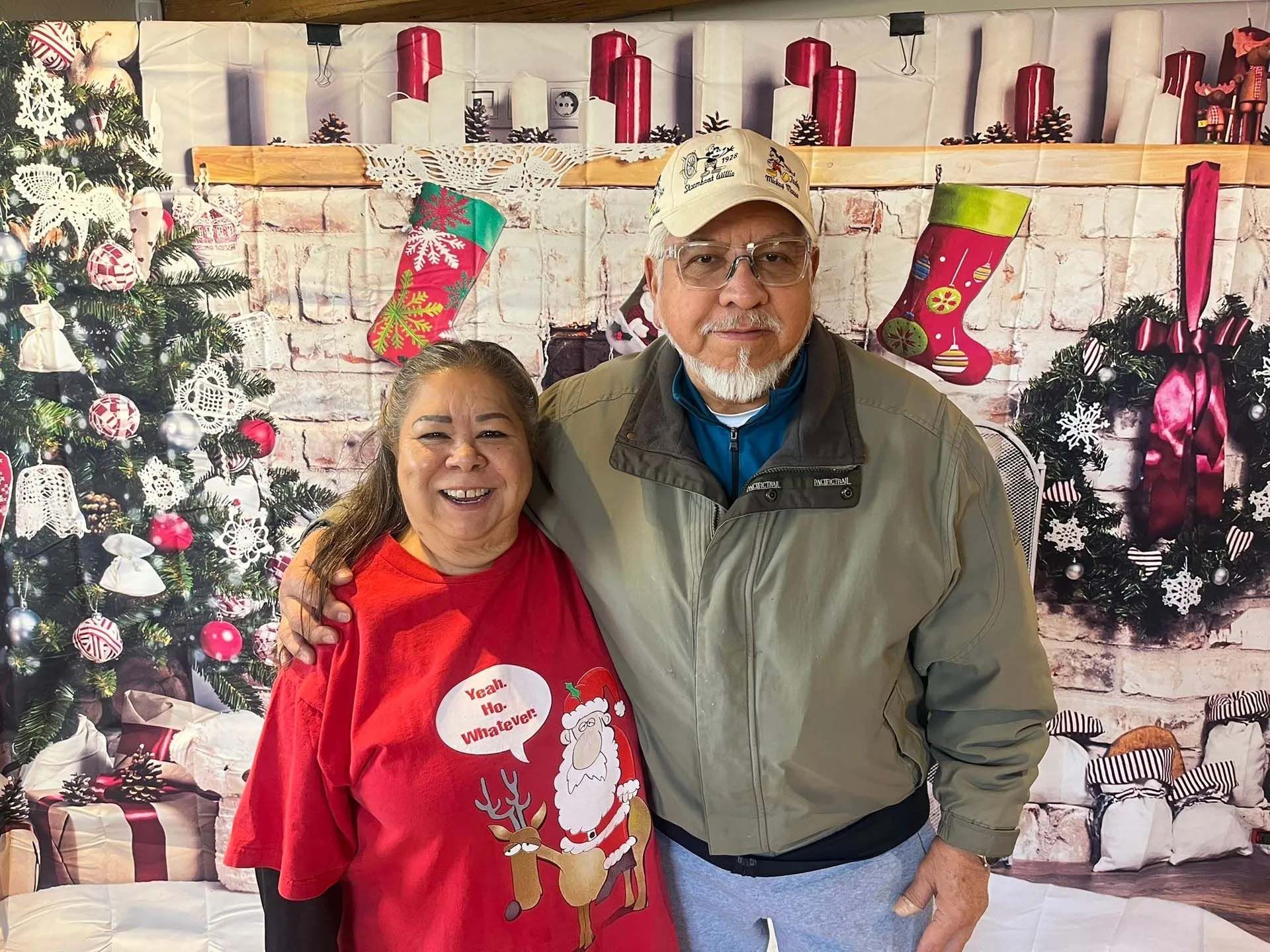 Couple smiling, posing in front of a Christmas backdrop with tree, stockings, and gifts.
