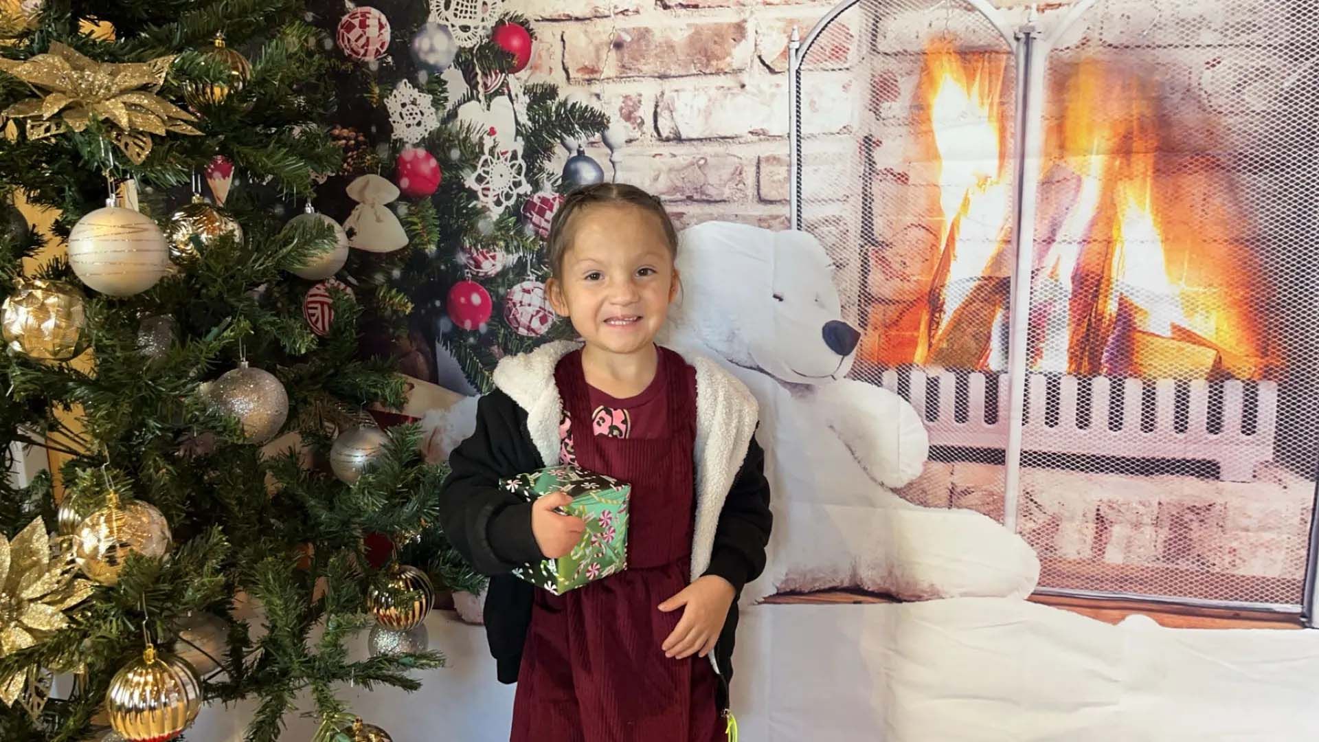 Girl holding present, smiling, in front of a Christmas tree and a fireplace backdrop.
