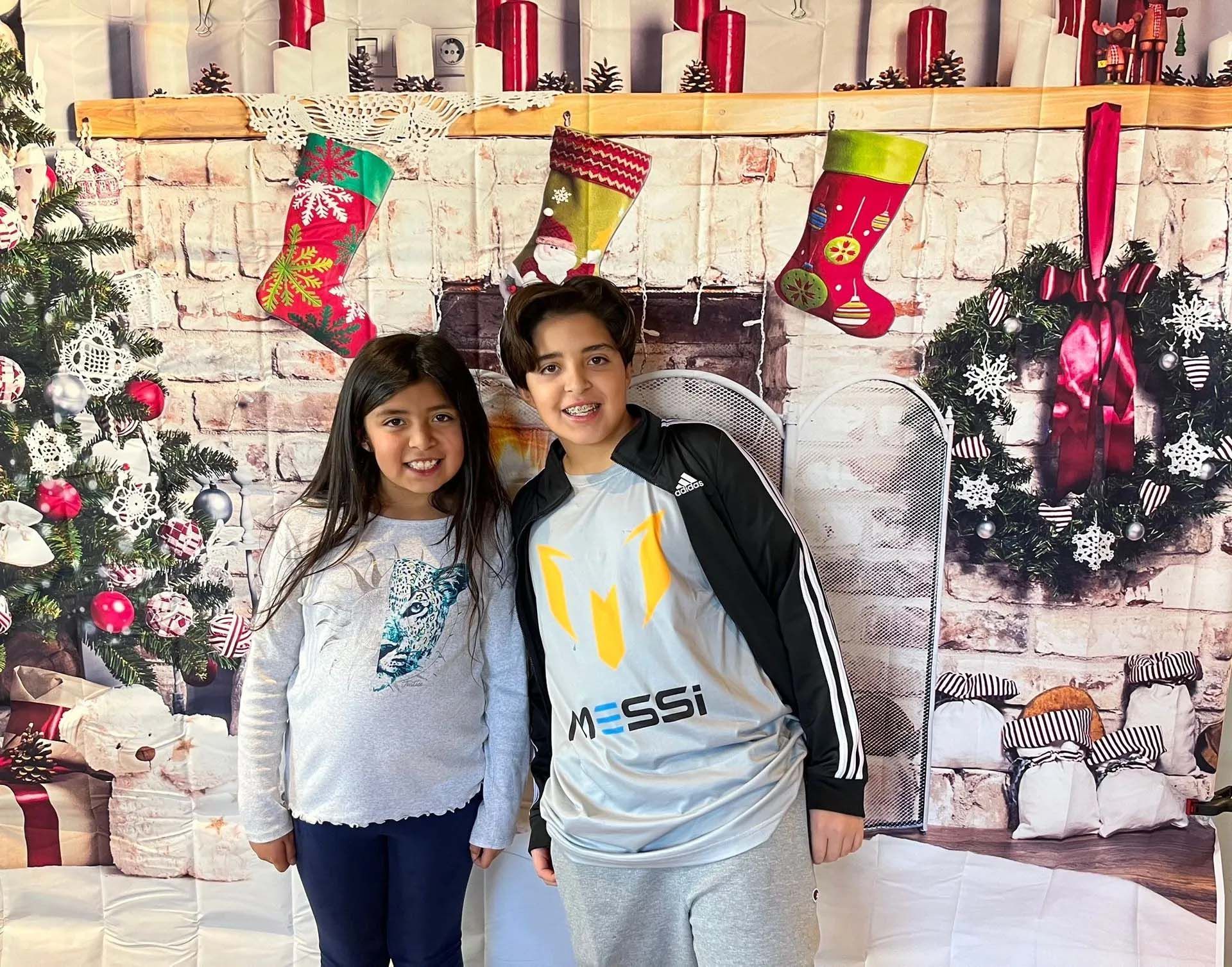 Two kids pose in front of a Christmas backdrop. Girl in sweater, boy in Messi shirt, smiling.