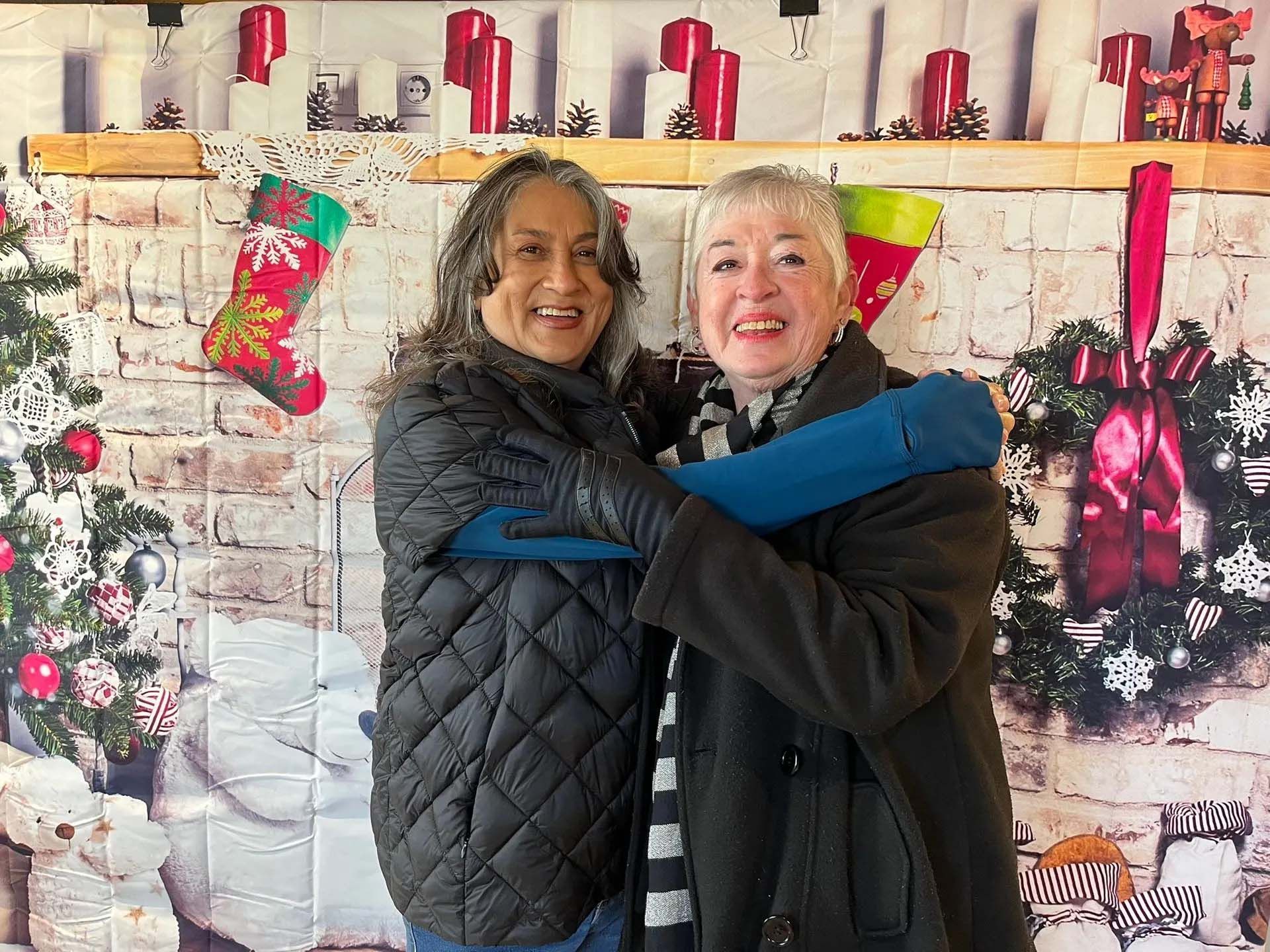 Two women embracing in front of a Christmas-themed backdrop. They smile, one in a black puffer jacket and the other in a black coat and scarf.