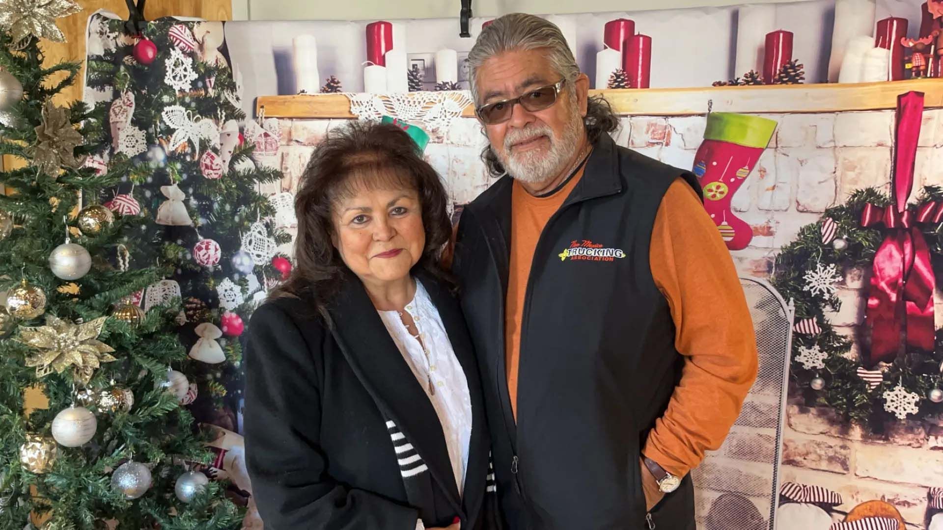 Couple smiling, posing in front of a Christmas-themed backdrop with a tree, garland and stockings.
