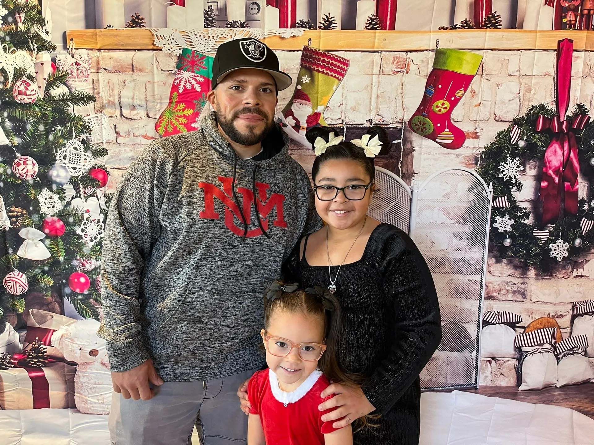 Family posing in front of a Christmas backdrop. Man in grey hoodie, two girls, one in red.