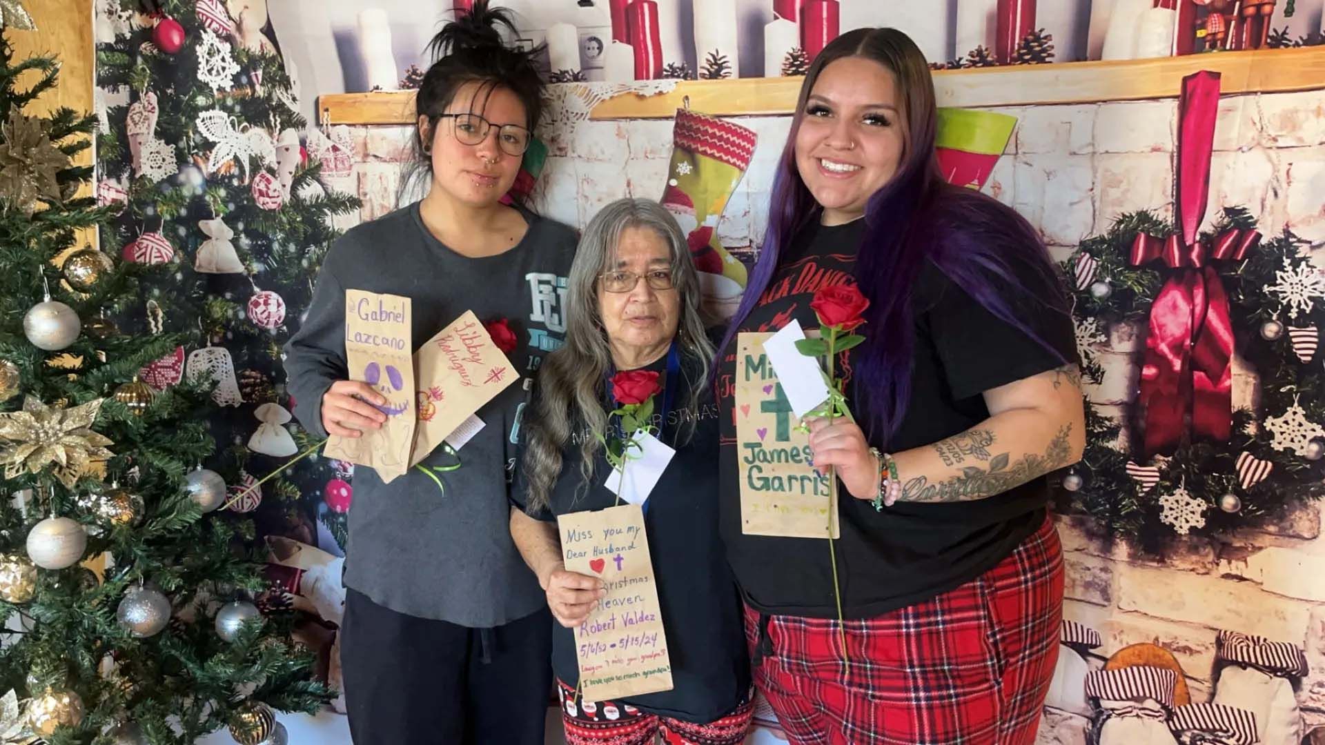 Three people holding cards and roses stand by Christmas decorations.