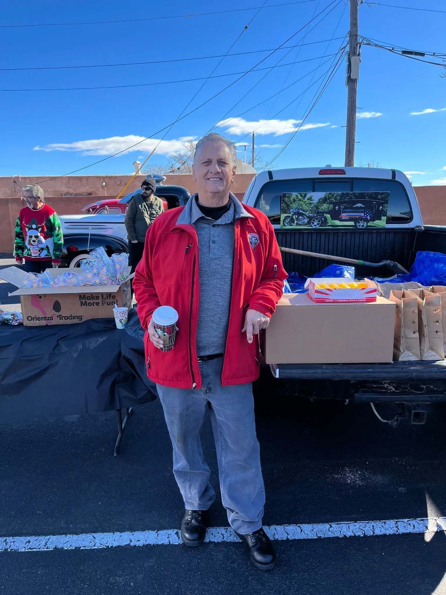 Man in red jacket stands by truck with donations, holding a mug, sunny day.