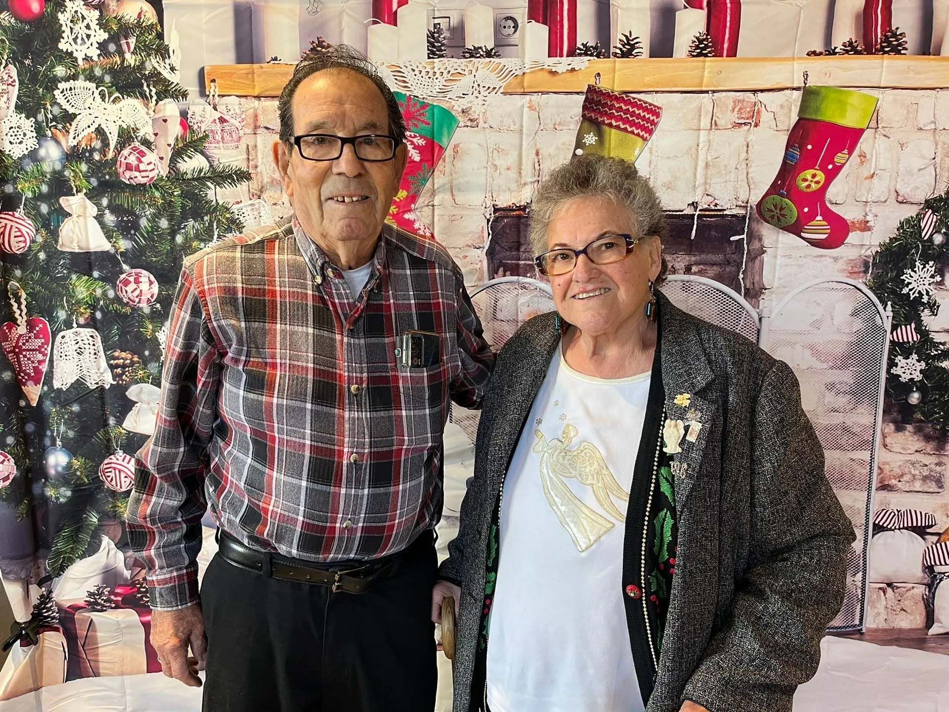 Couple posing in front of a Christmas backdrop. Man in plaid shirt, woman in black jacket, both wearing glasses.