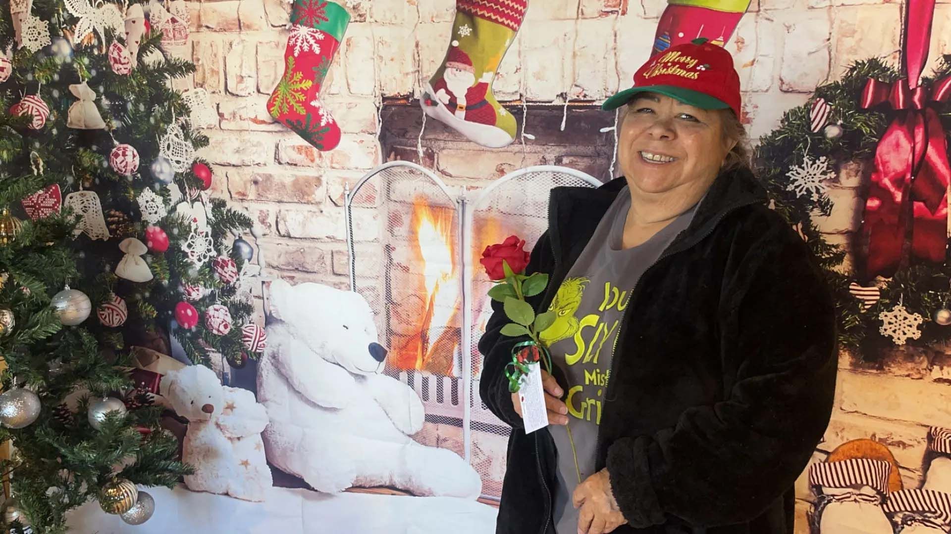Woman in a Christmas hat holding a rose, posed in front of a holiday backdrop with a Christmas tree.
