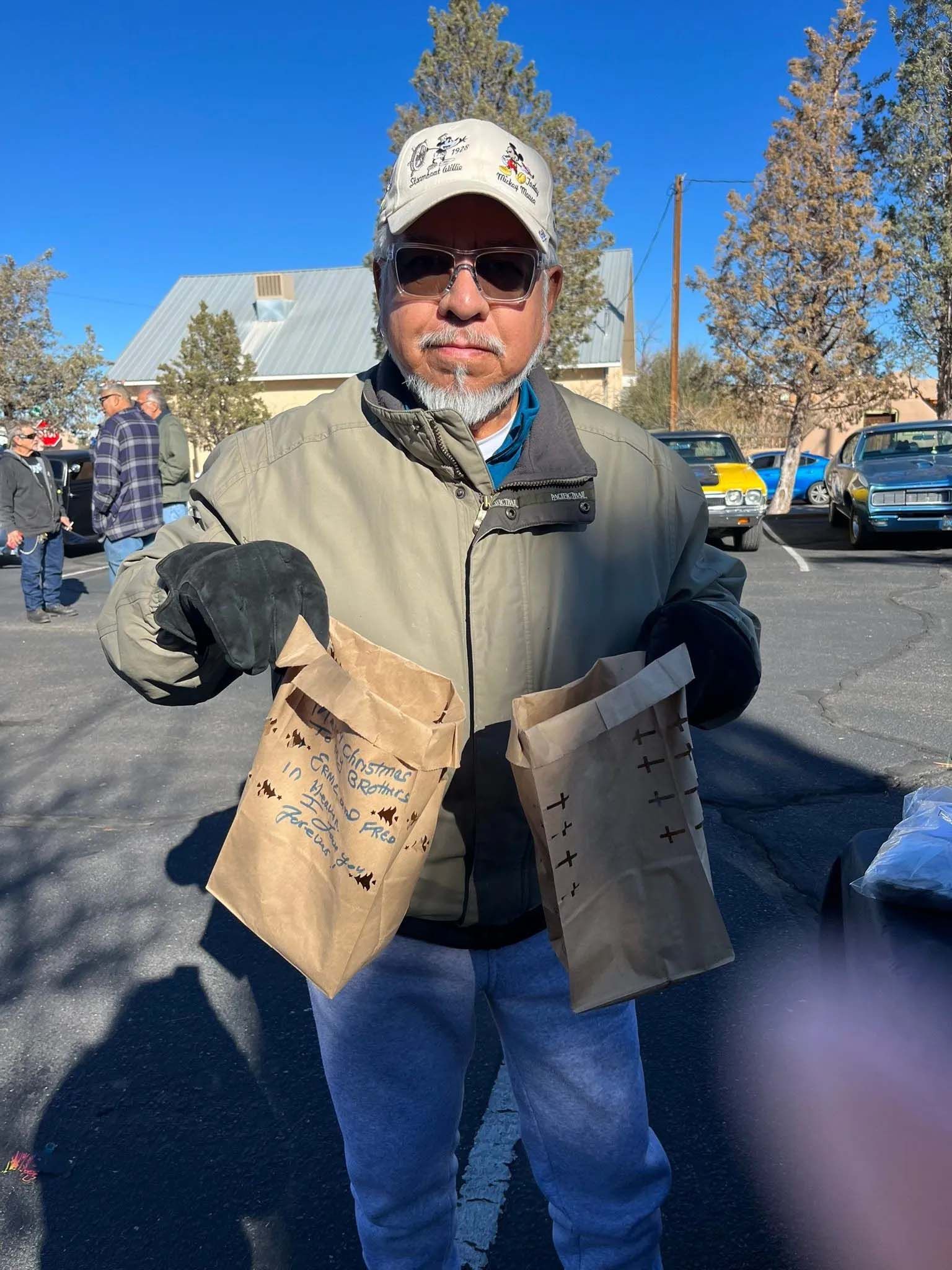 Man holding two brown paper bags, outdoors in a parking lot. He is wearing a jacket, hat, and sunglasses.