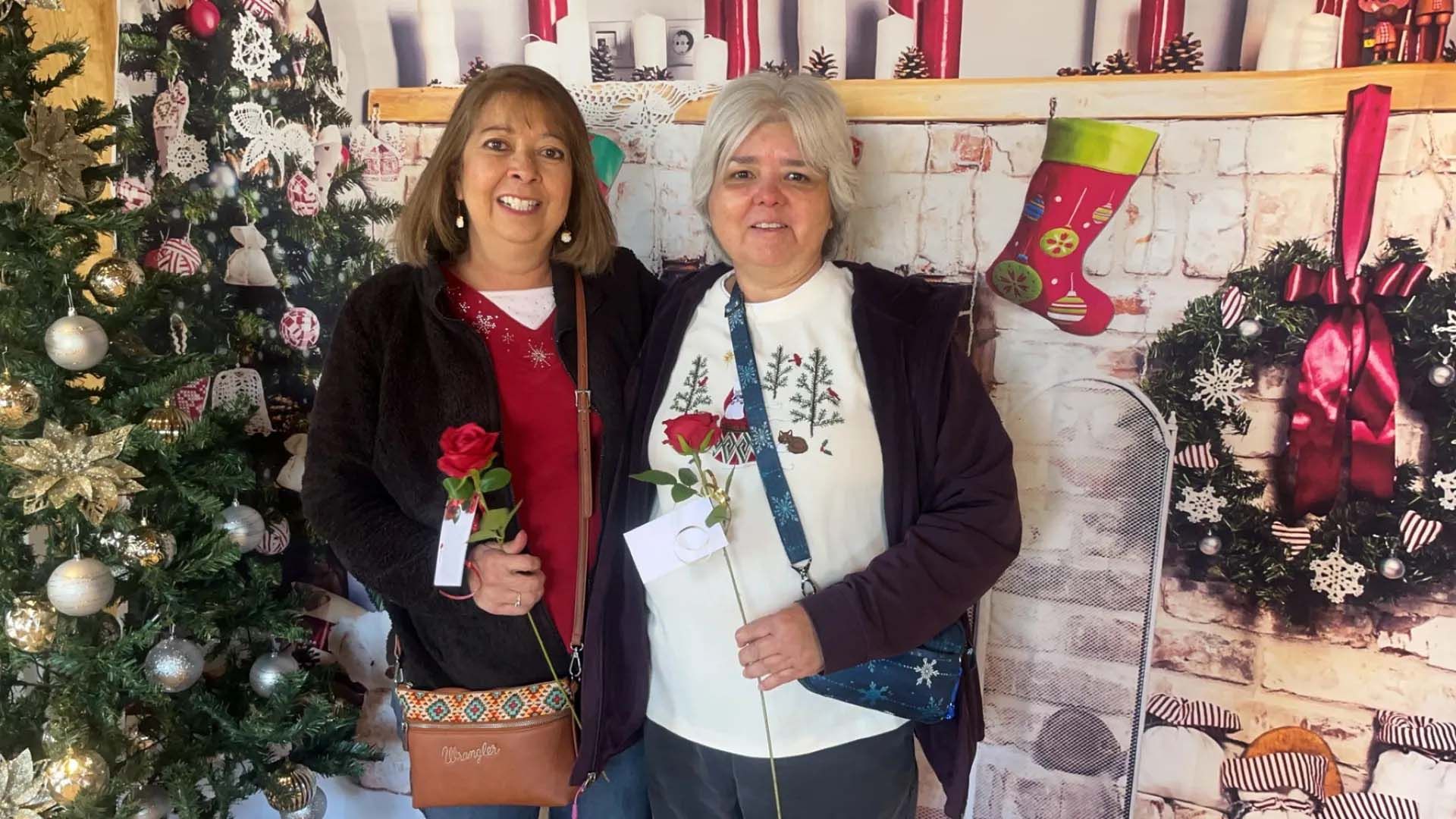 Two women pose with red roses in front of a Christmas backdrop.