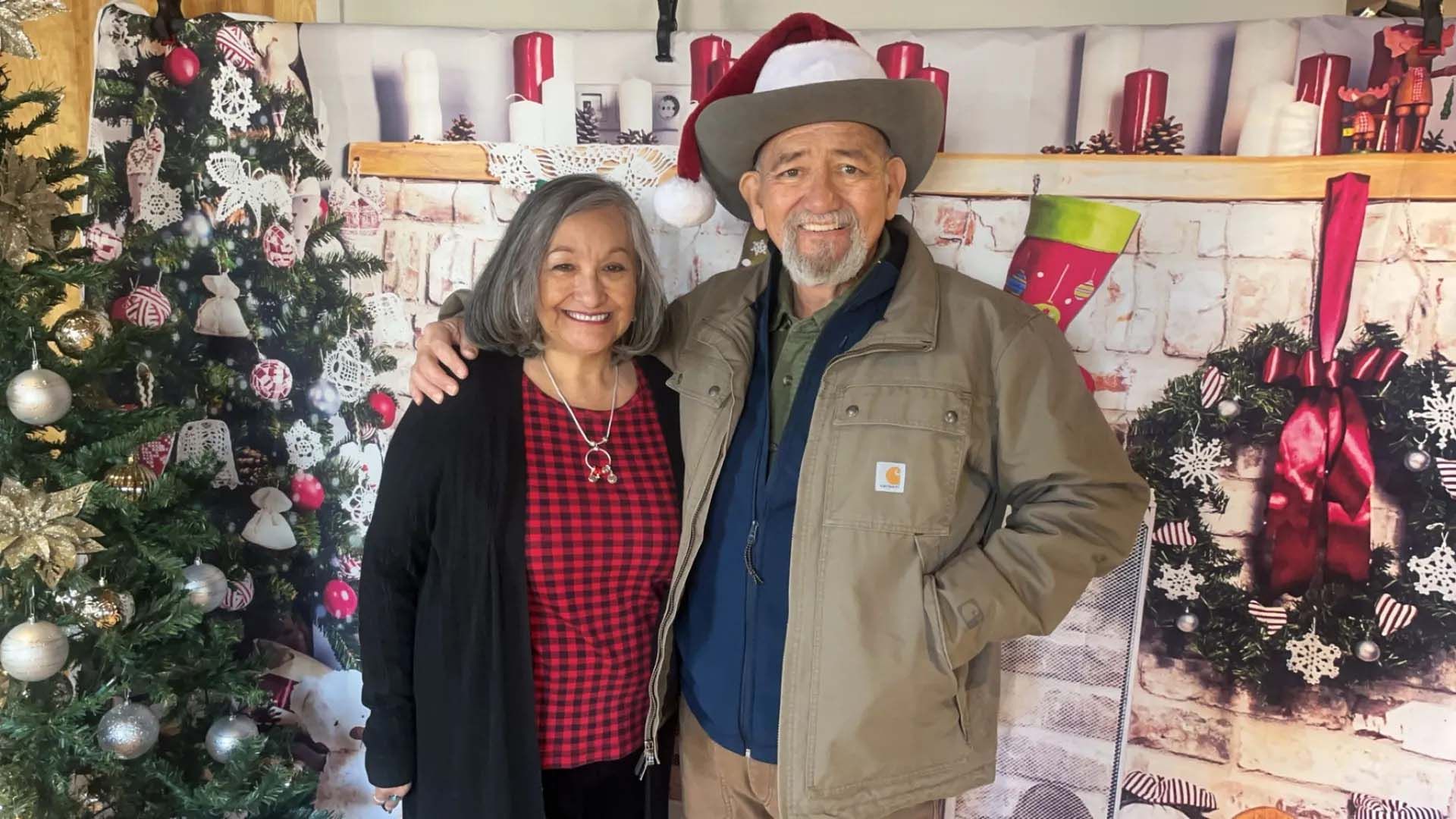 Couple posing in front of a Christmas tree and fireplace backdrop. Man wearing a Santa hat and work jacket, woman in a red shirt.