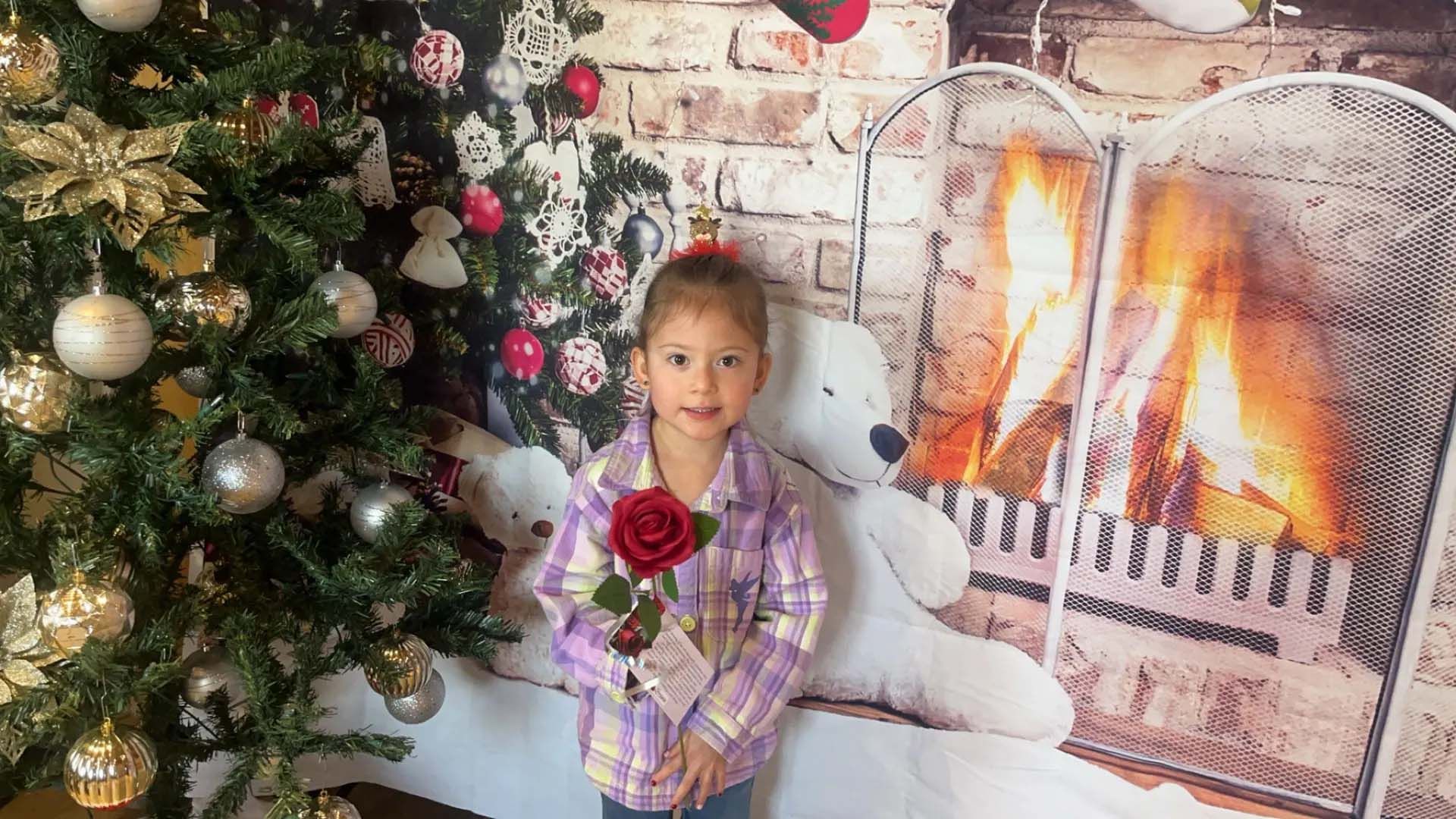 Girl in plaid shirt with a rose stands in front of a Christmas tree, fireplace backdrop, and teddy bear.
