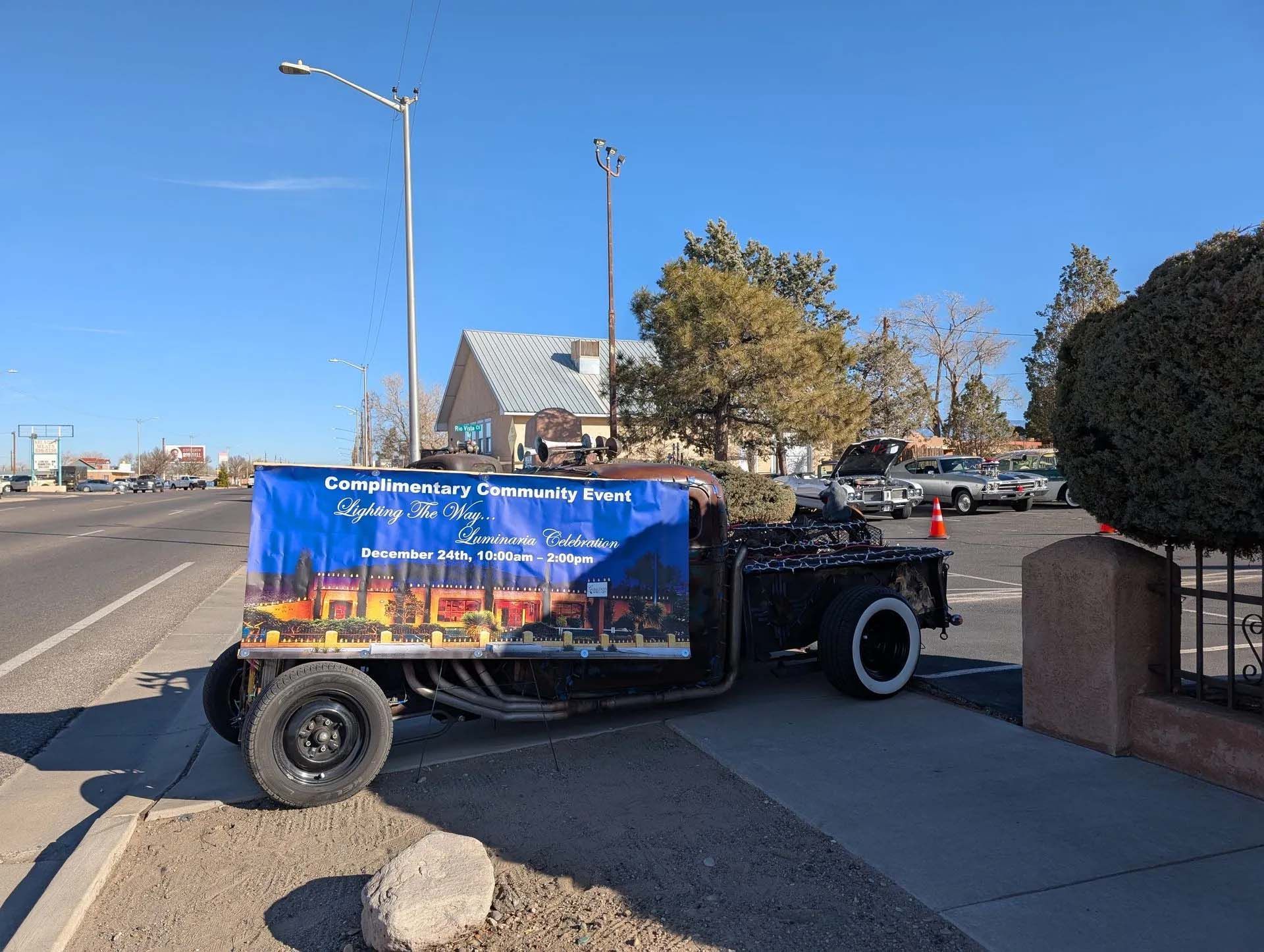 Black hot rod truck with a blue banner parked by a sidewalk in front of buildings on a sunny day.