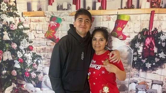 A young person in black hoodie and a woman in red shirt pose in front of a Christmas backdrop, embracing.