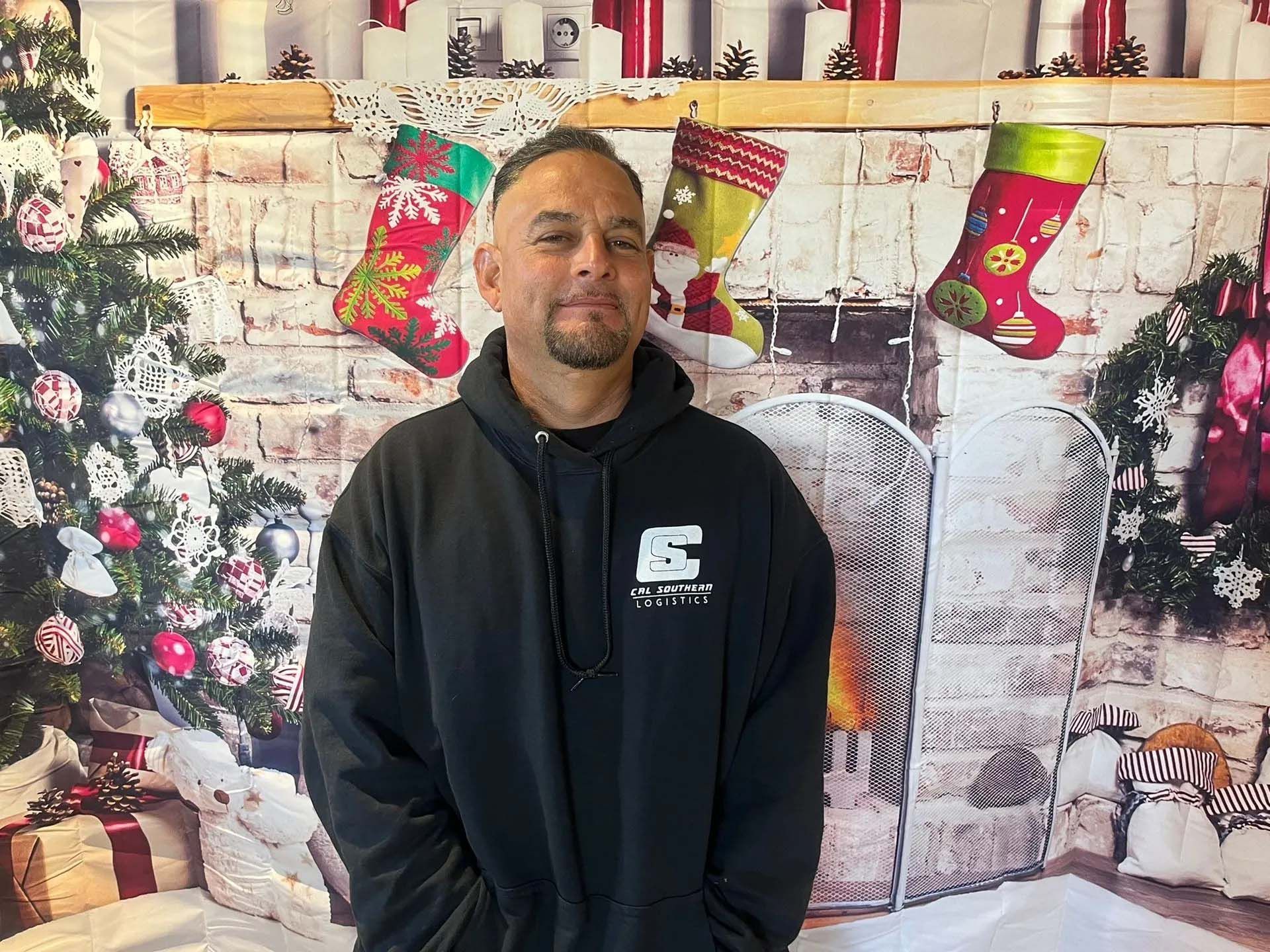 Man in black hoodie stands in front of a Christmas-themed backdrop with a fireplace, stockings, and a decorated tree.